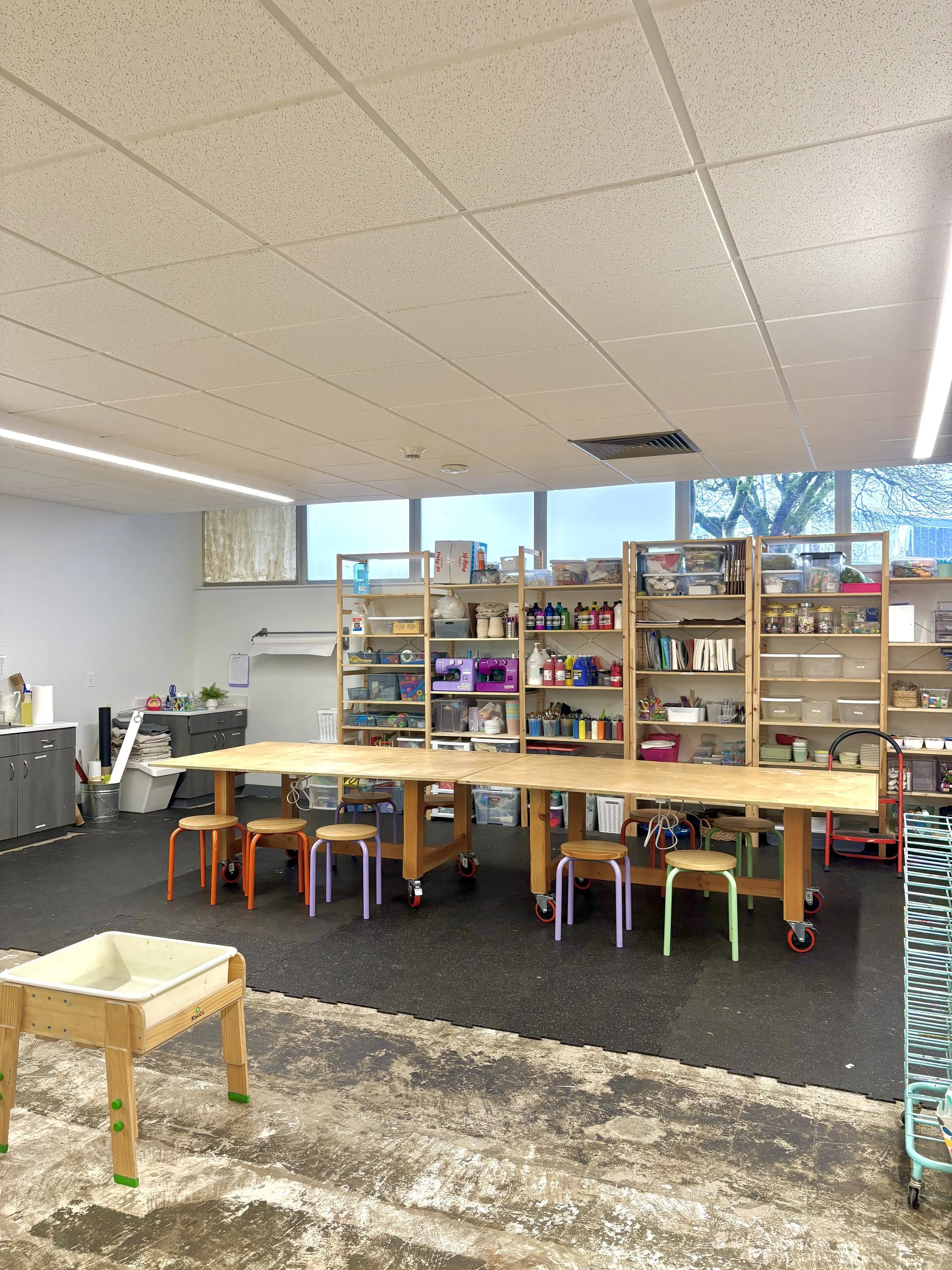 A spacious classroom or craft room with a long wooden table on wheels, surrounded by colorful stools. Shelves filled with art supplies, books, and bins line the wall. The room has large windows letting in natural light.