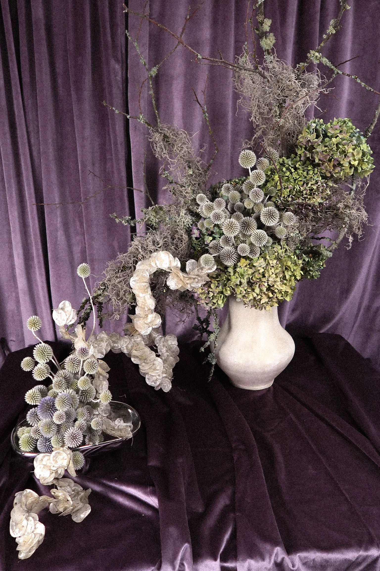 A large white vase with dried flowers, branches, and moss, placed on a black fabric surface with a purple cloth backdrop.
