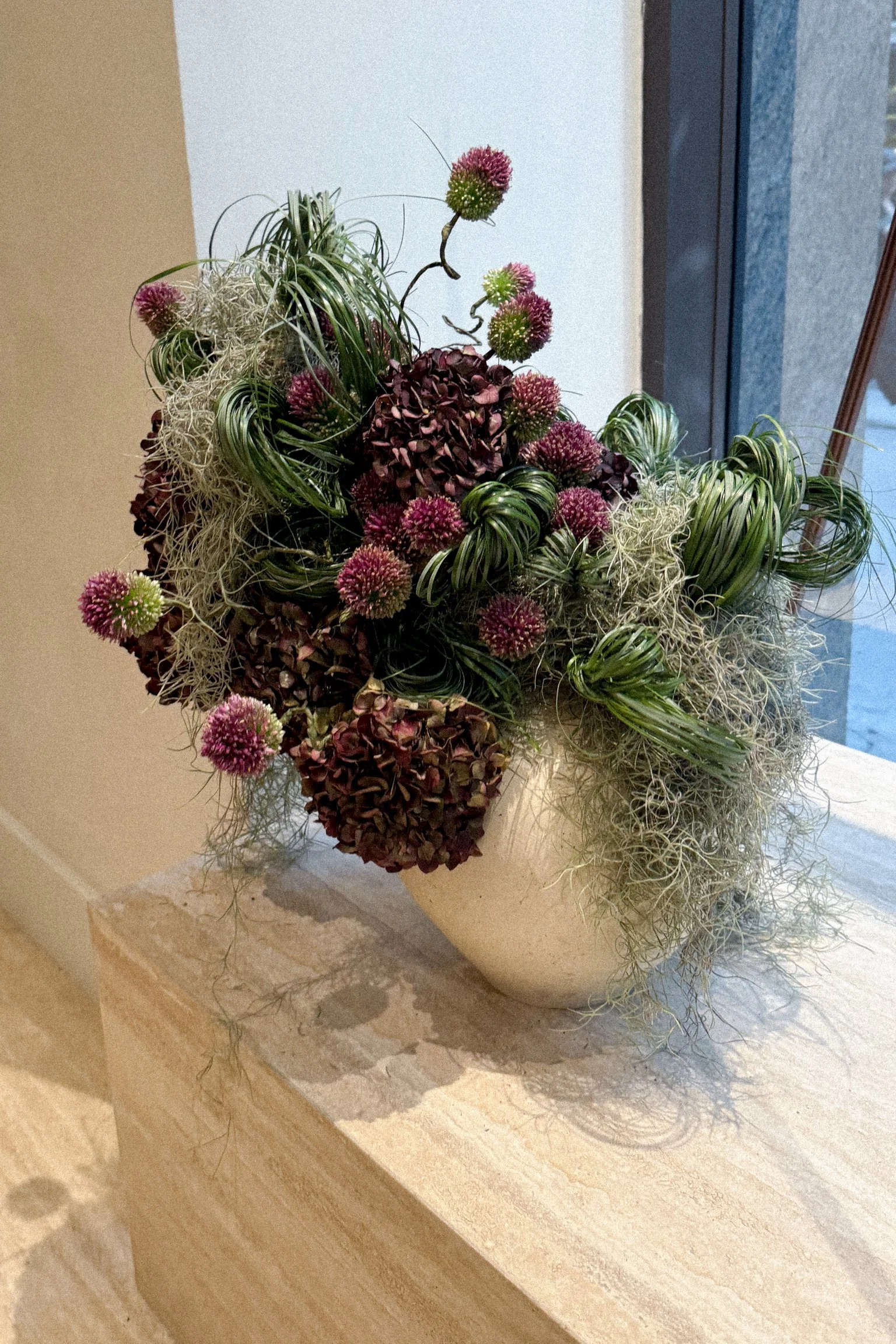 A floral arrangement featuring pink and burgundy flowers, curly green leaves, and white, wispy Spanish moss in a white ceramic vase on a beige marble surface near a window.