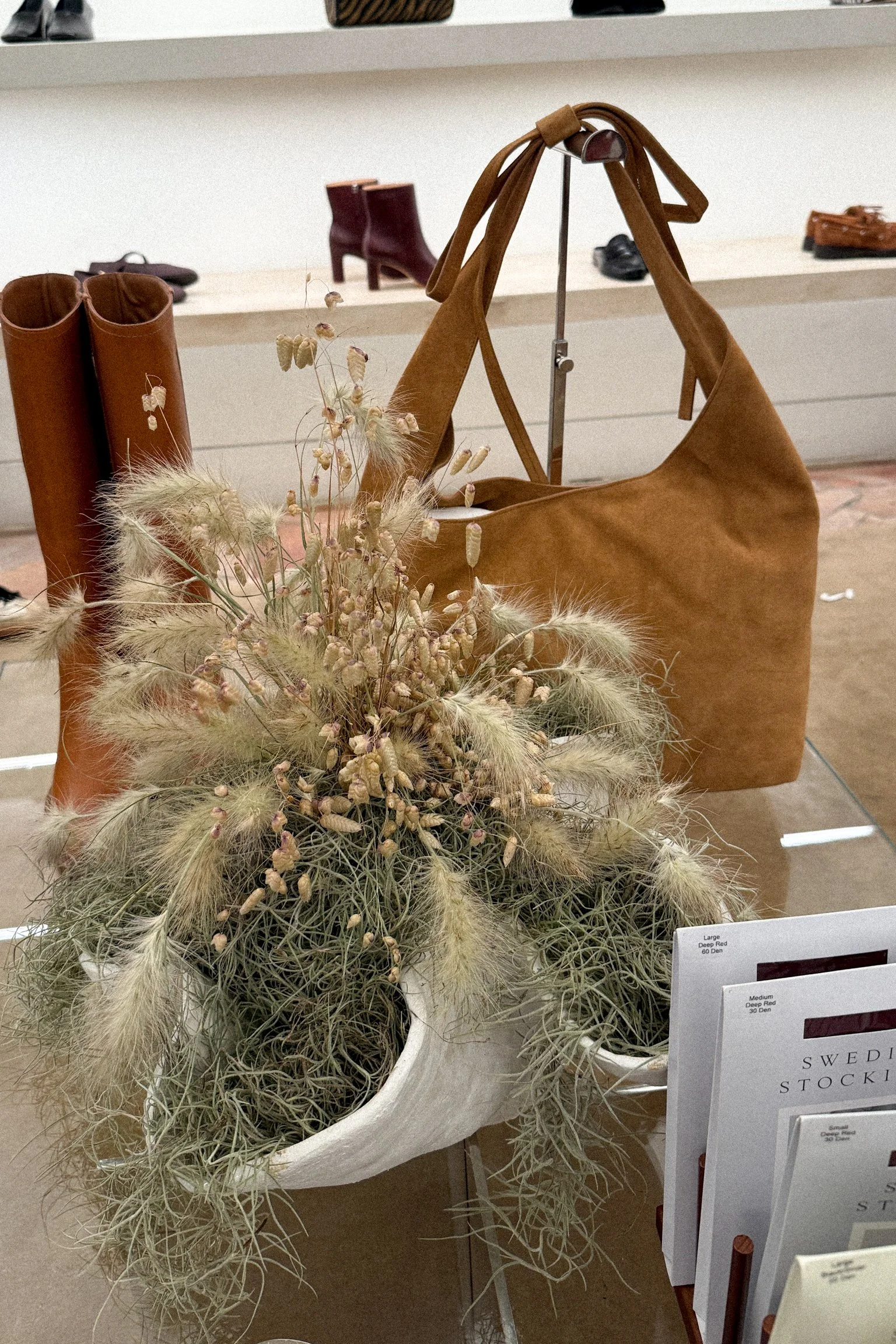 Display of a large tan leather shoulder bag, a pair of brown leather boots, and dried flowers in a white bowl, within a store setting.