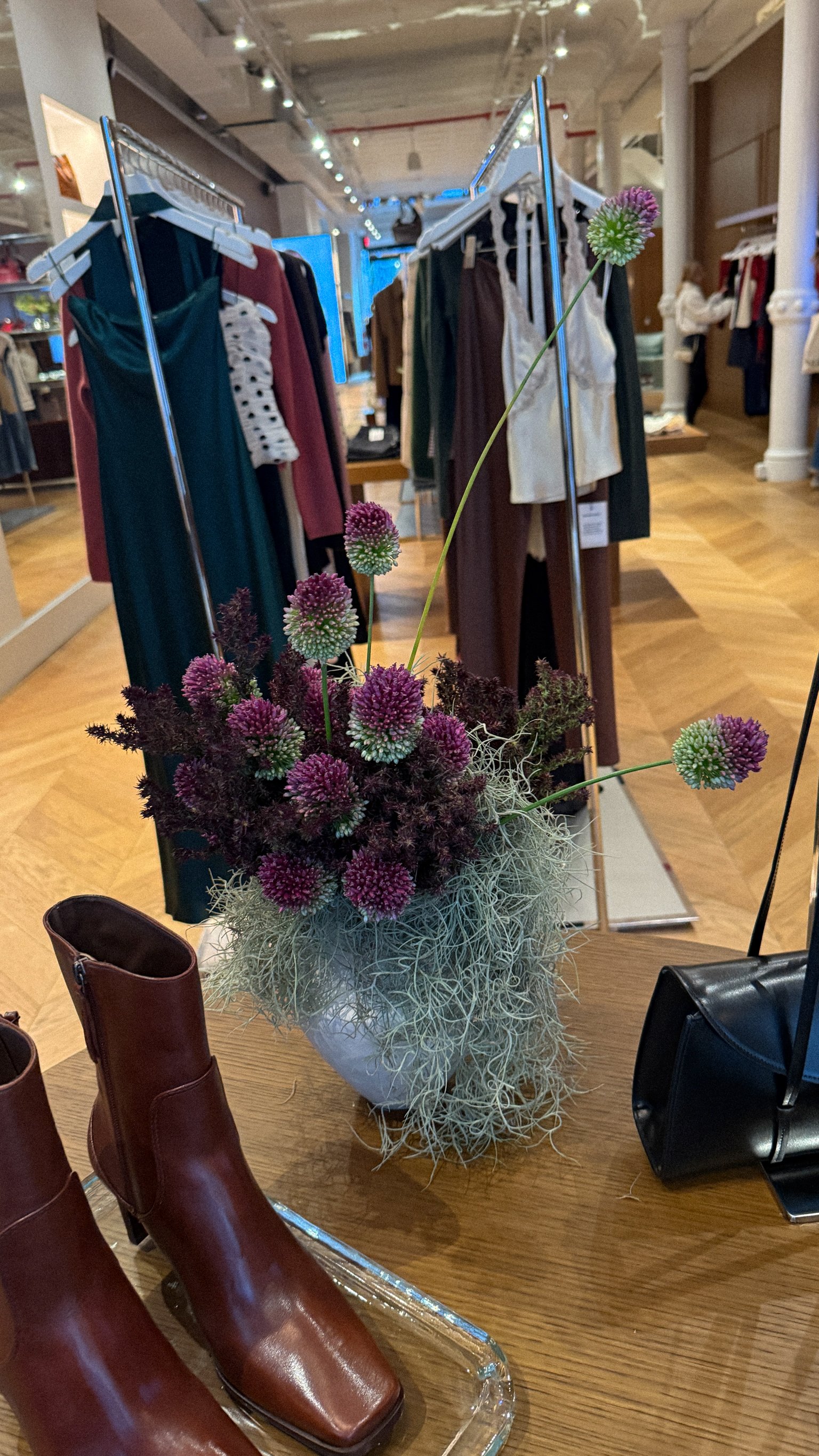 A floral arrangement with purple and white globe amaranth flowers and grayish-green air plants on a wooden table in a clothing store display.