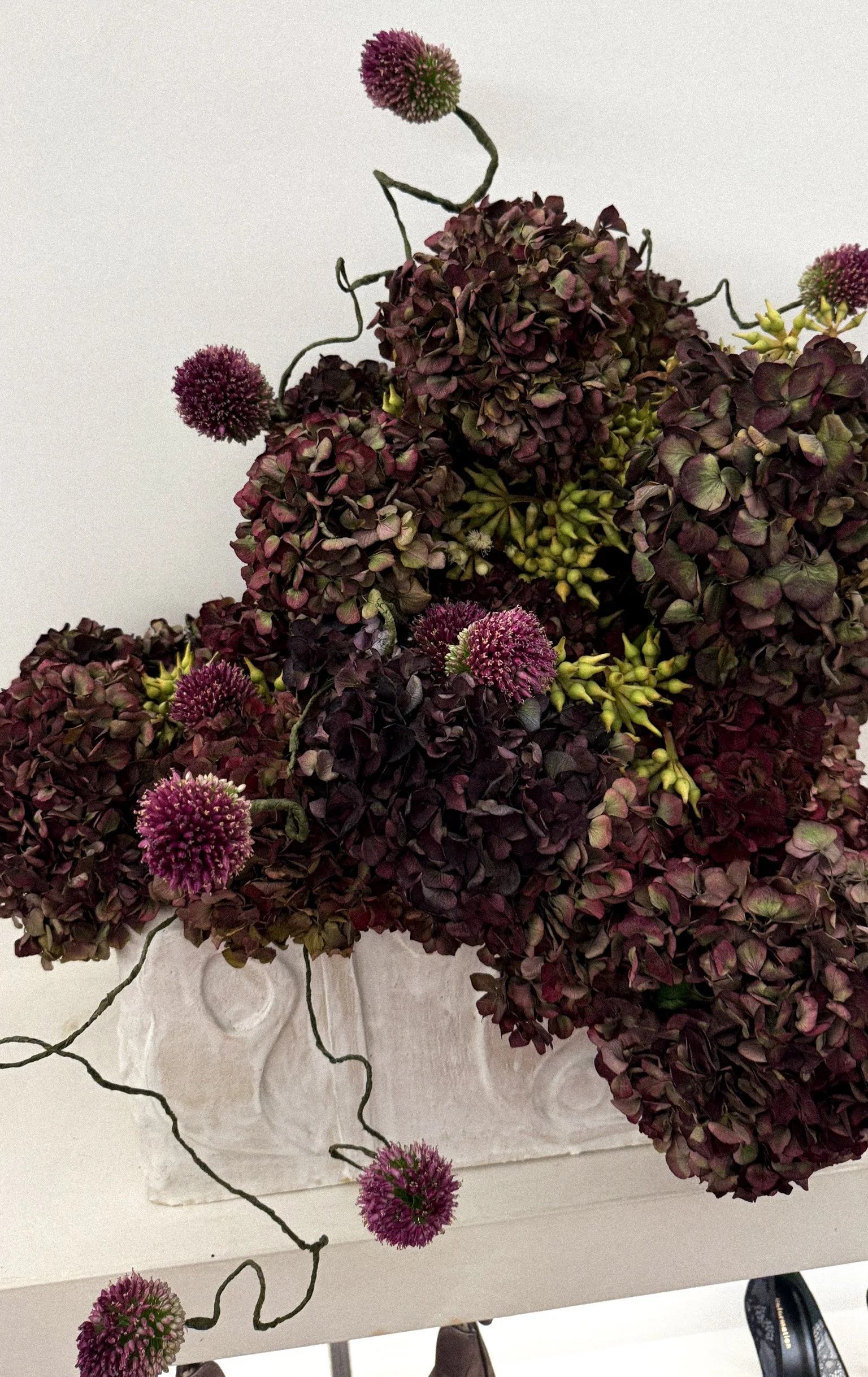 A bouquet of dark purple hydrangeas and round purple allium flowers in a white textured vase.