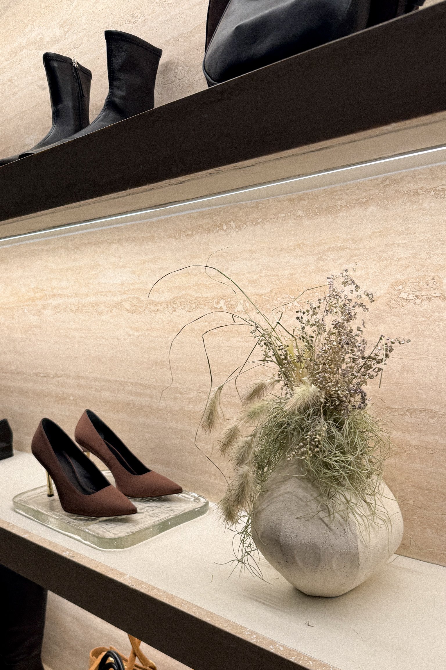 Display of black boots on the top shelf, brown high heels and a vase with dried flowers on the bottom shelf in a store.