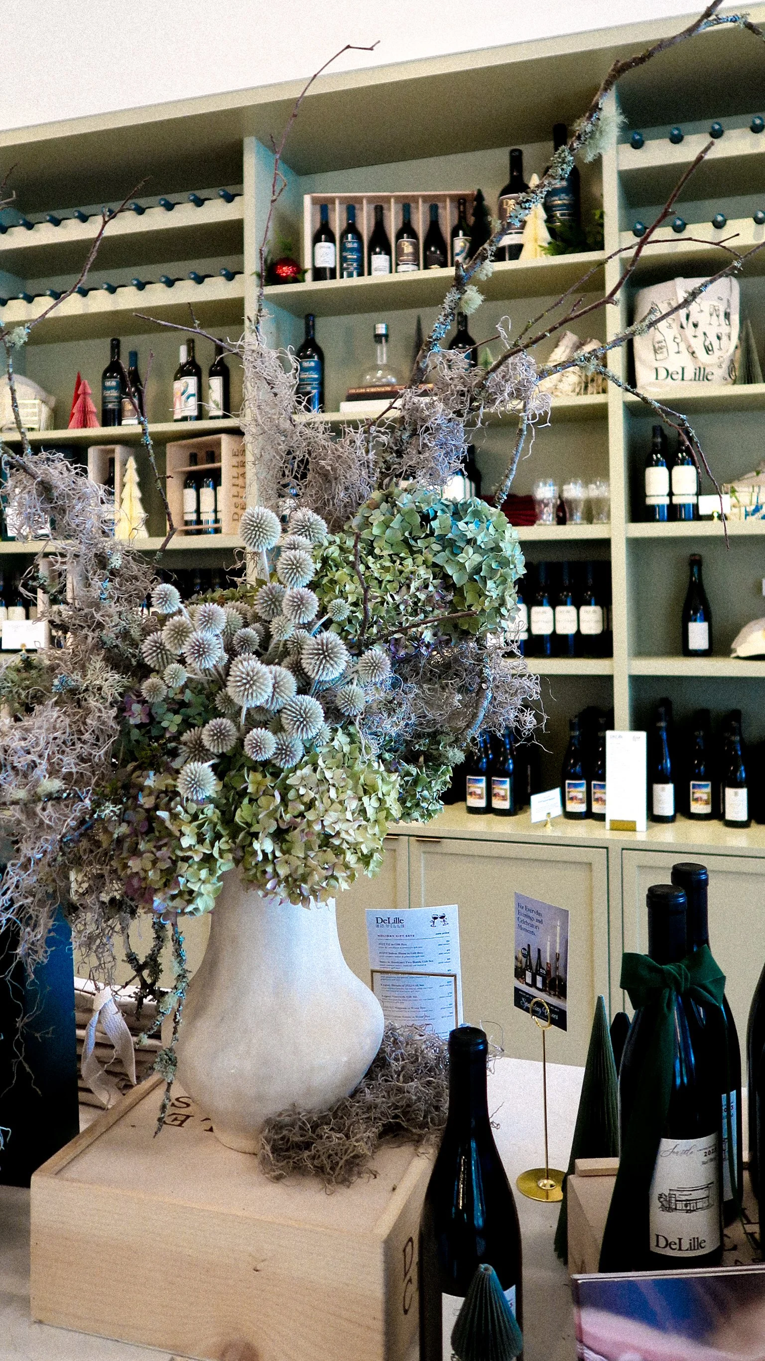 A large beige ceramic vase filled with dried flowers, branches, and succulents, placed on a wooden box in a store or winery with shelves of wine bottles in the background.
