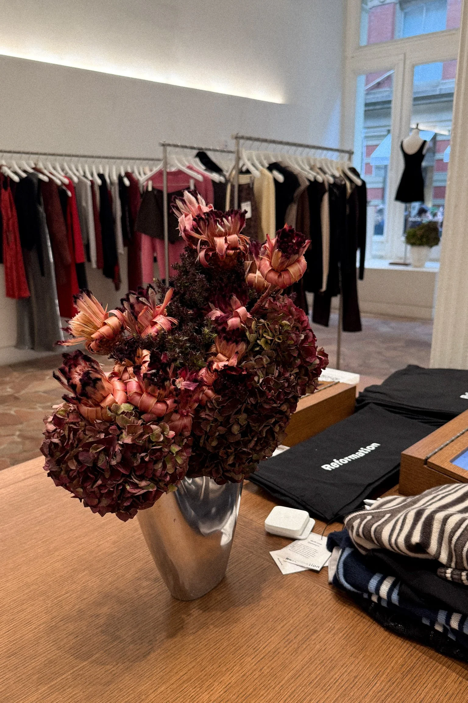 A vase with dried flowers on a wooden table in a clothing store, with racks of clothes in the background and a window with natural light.