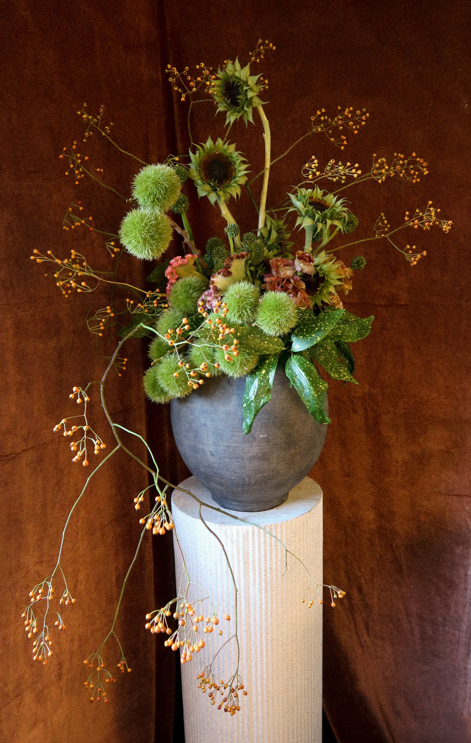 A flower arrangement with sunflowers, green fluffy balls, and orange accents in a dark gray vase on a white pedestal against a dark wooden background.