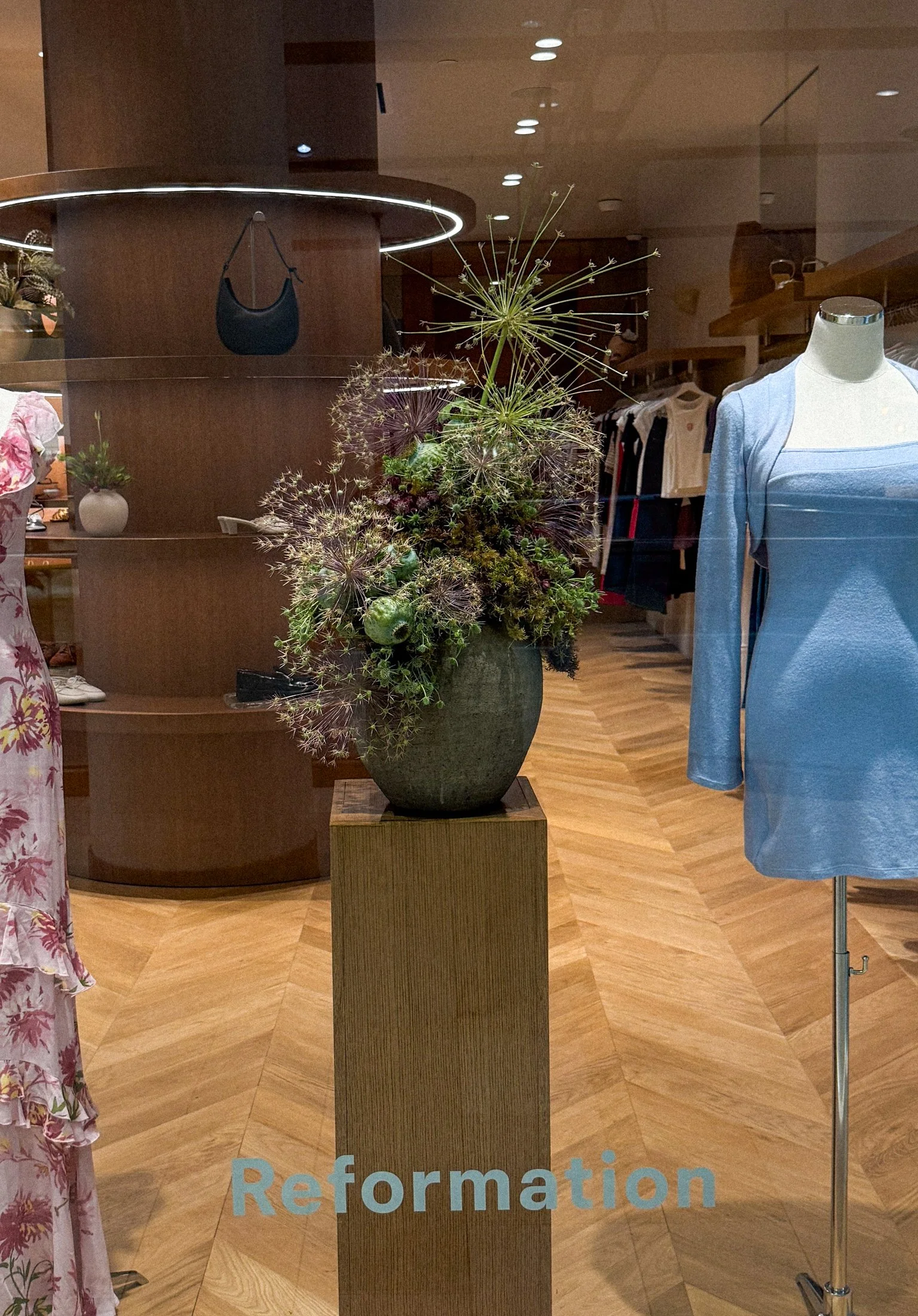 A floral arrangement in a green pot on a wooden pedestal inside a retail store, with clothing displays and racks of garments in the background.