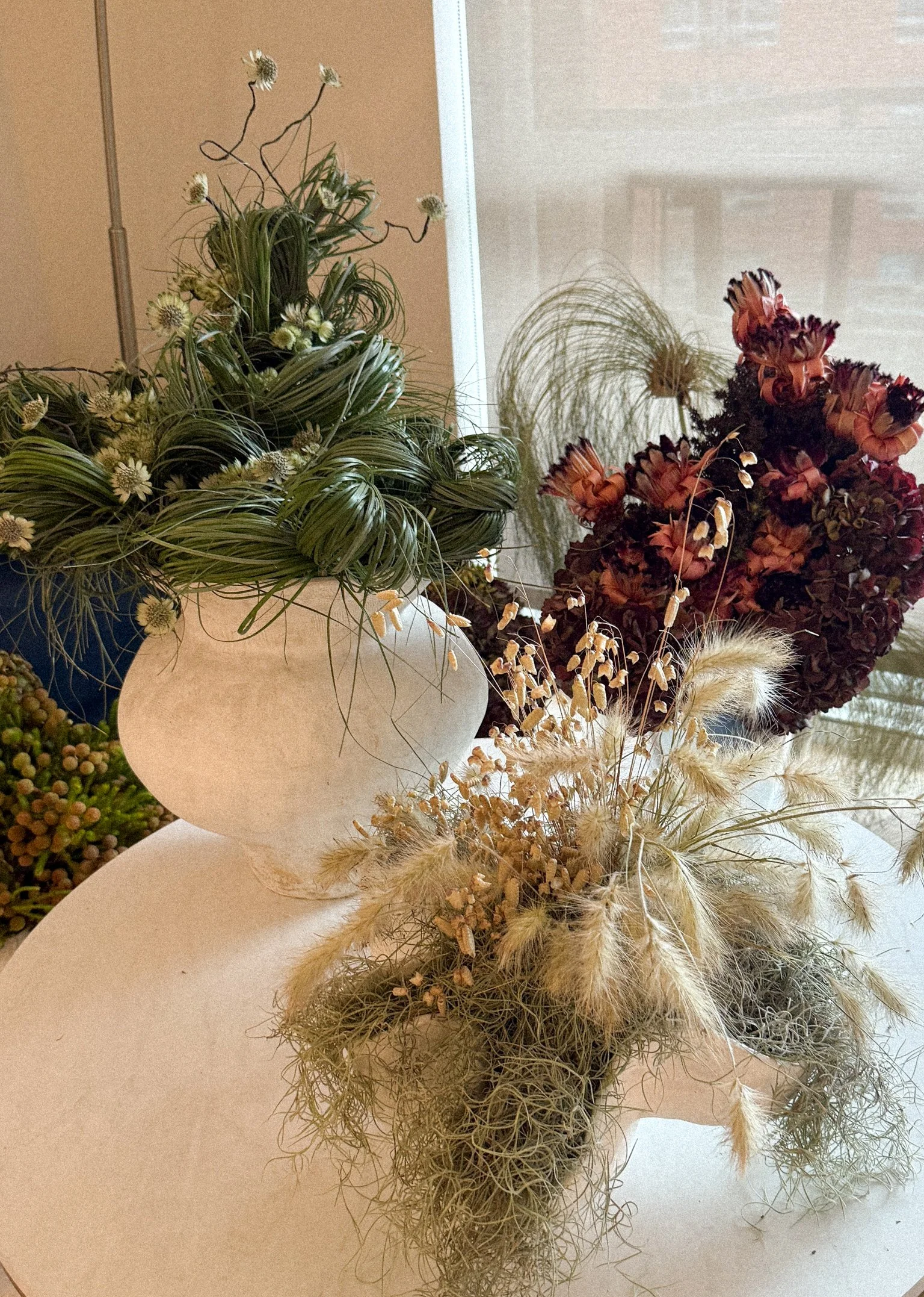 Arrangement of dried flowers and greenery in a white ceramic vase on a white surface, with a backdrop of large window and beige wall.