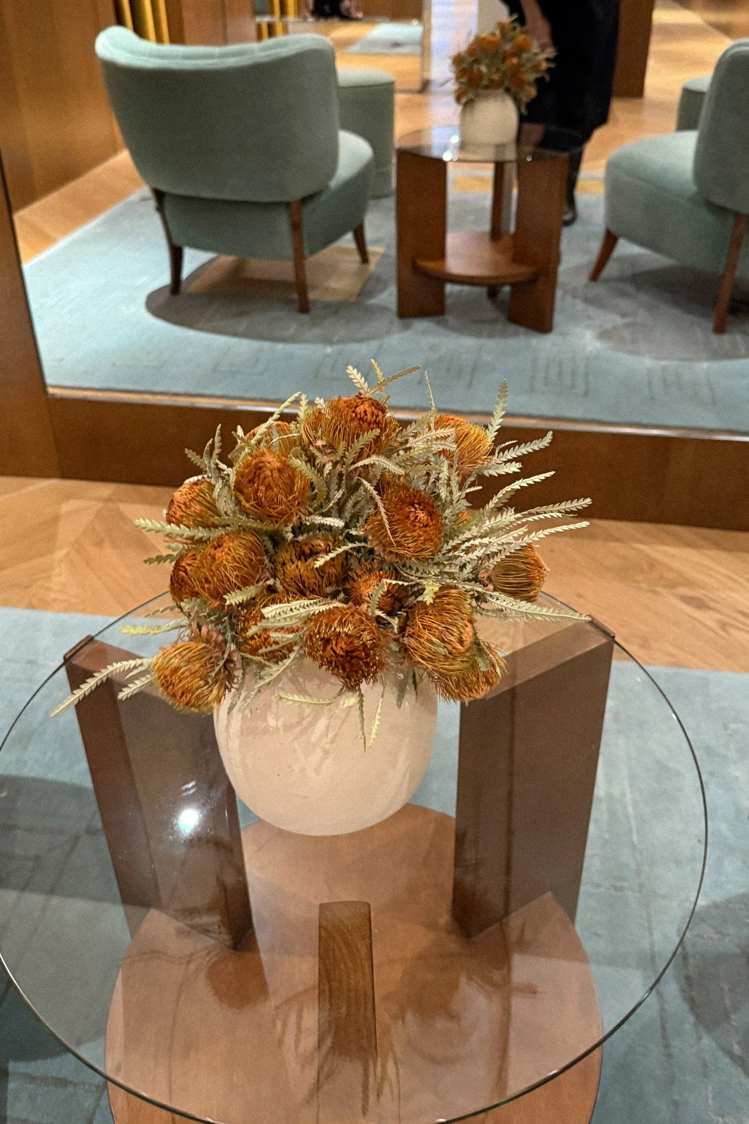 A decorative ceramic vase with dried flowers placed on a glass-top wooden table in a lobby or waiting area.