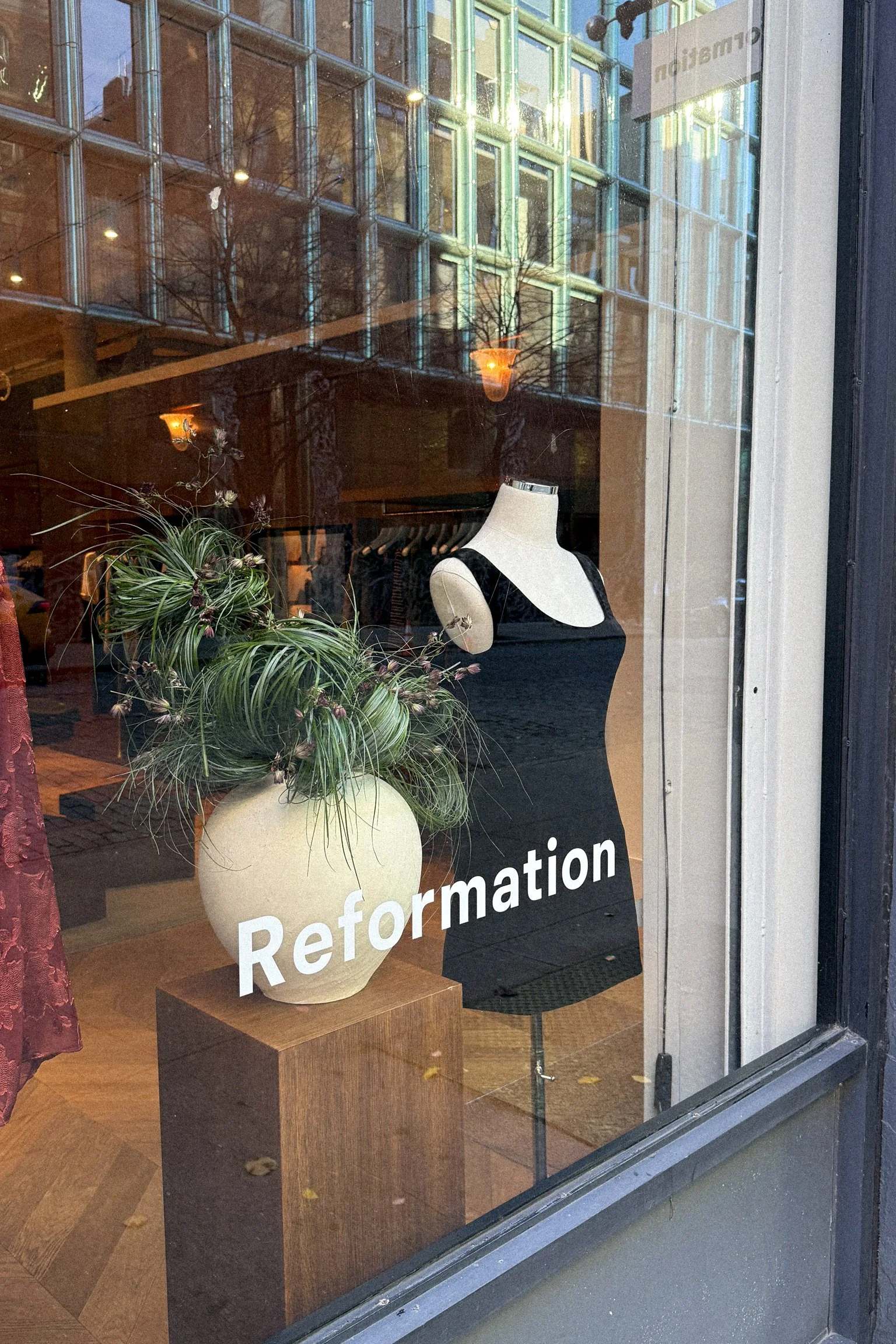 A storefront window display with a sign that reads 'Reformation'. Inside, there is a mannequin dressed in a black dress, and a large vase with green plants and flowers on a wooden pedestal.