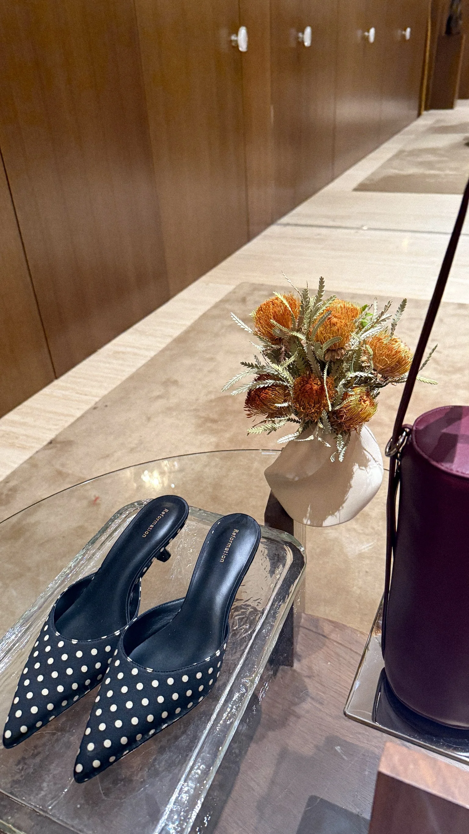 Polka dot high heels, a vase with orange flowers, purple bag, and a glass table with wooden background.