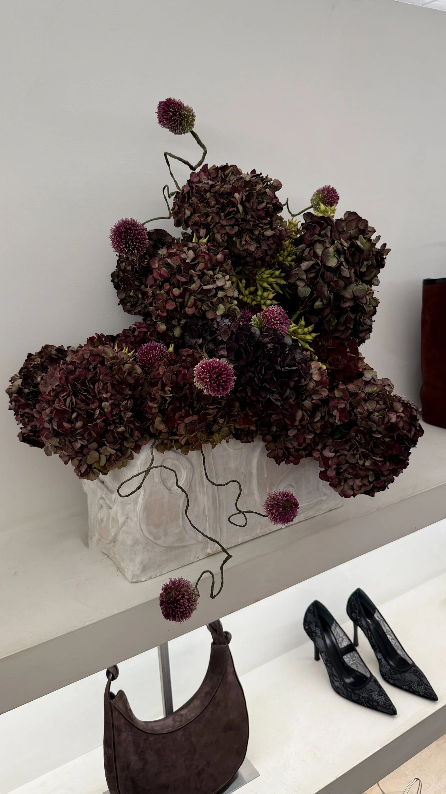 A white vase containing dark purple hydrangeas and pink pom-pom flowers placed on a shelf. Below the shelf, black high-heeled shoes with lace detail and a brown handbag are visible.