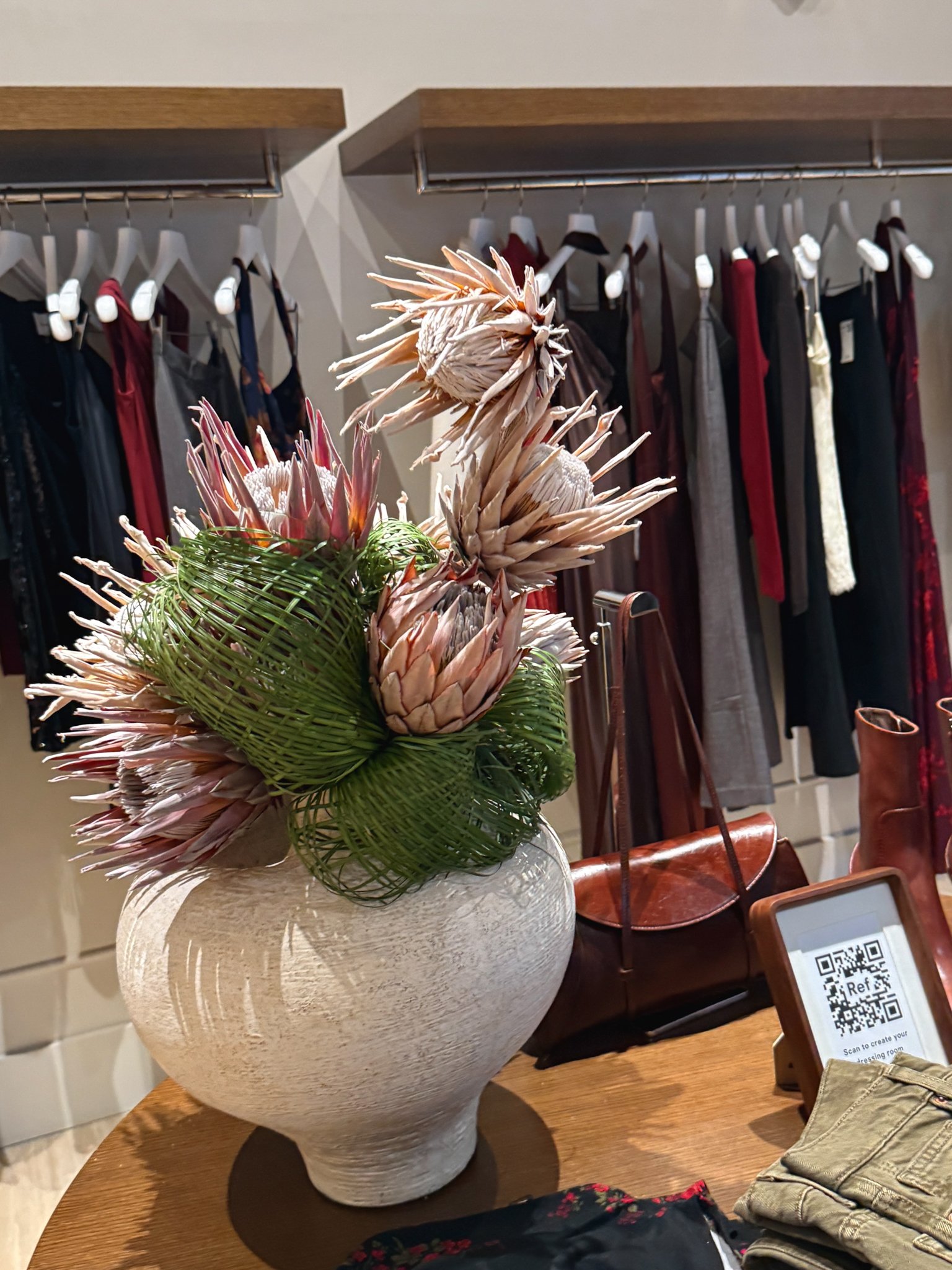 A white ceramic vase with large, pink protea flowers and green leaves on a wooden table inside a store, with clothing racks and a shopping bag in the background.