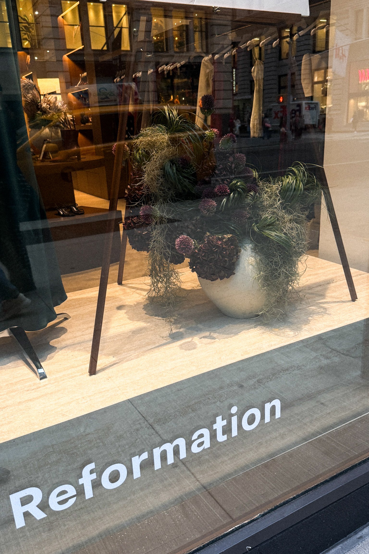A store window display featuring a large marble vase with purple and green plants, placed on a wooden surface. The word 'Reformation' is printed on the glass at the bottom of the display.