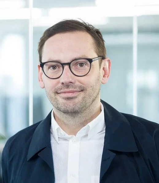 Portrait von Martin Jordan – mit kurzen Haaren, Brille in einem weißen Hemd in einer Büroumgebung