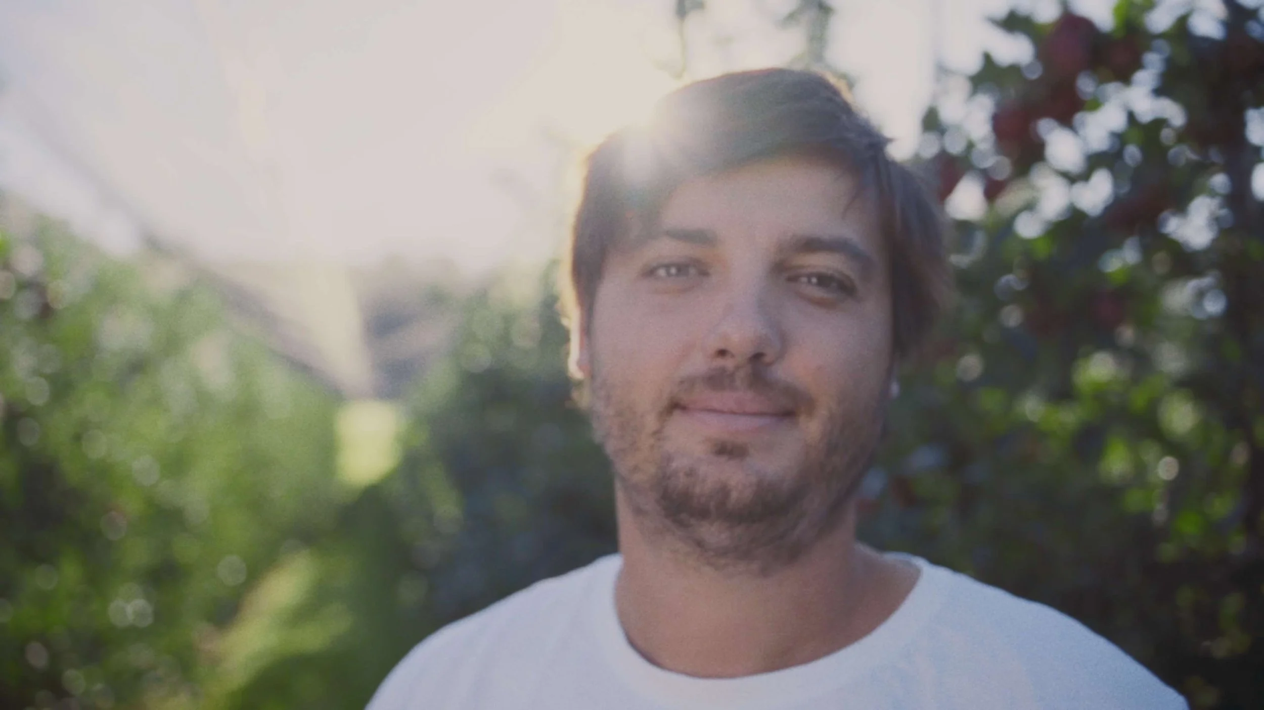 A man with dark hair and a beard smiling outdoors with sunlight behind him, trees and greenery in the background.