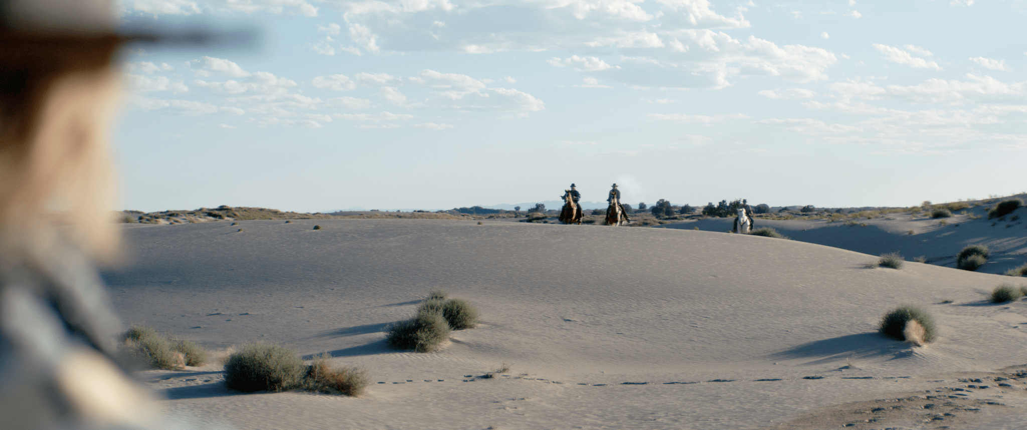 A person in the foreground partially out of focus looking towards three people on horseback riding across sandy desert dunes with sparse vegetation under a partly cloudy sky.