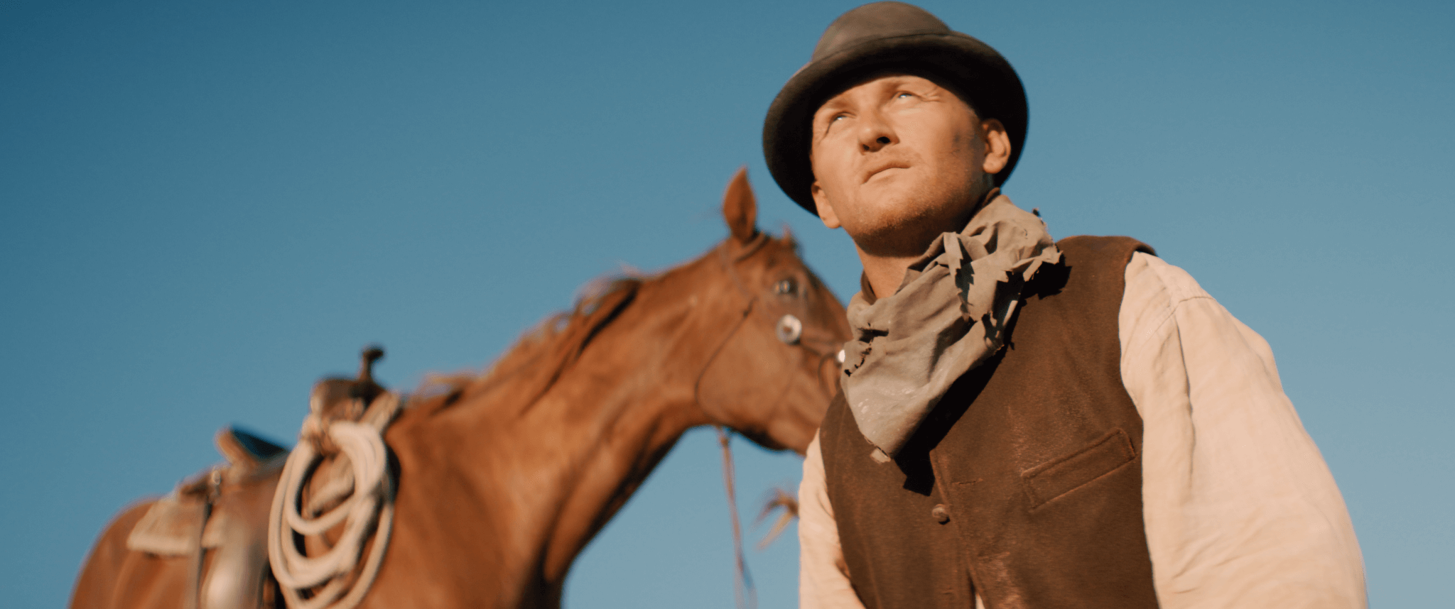 A man dressed as a cowboy with a brown vest, beige shirt, and bandana around his neck, standing next to a brown horse against a clear blue sky.