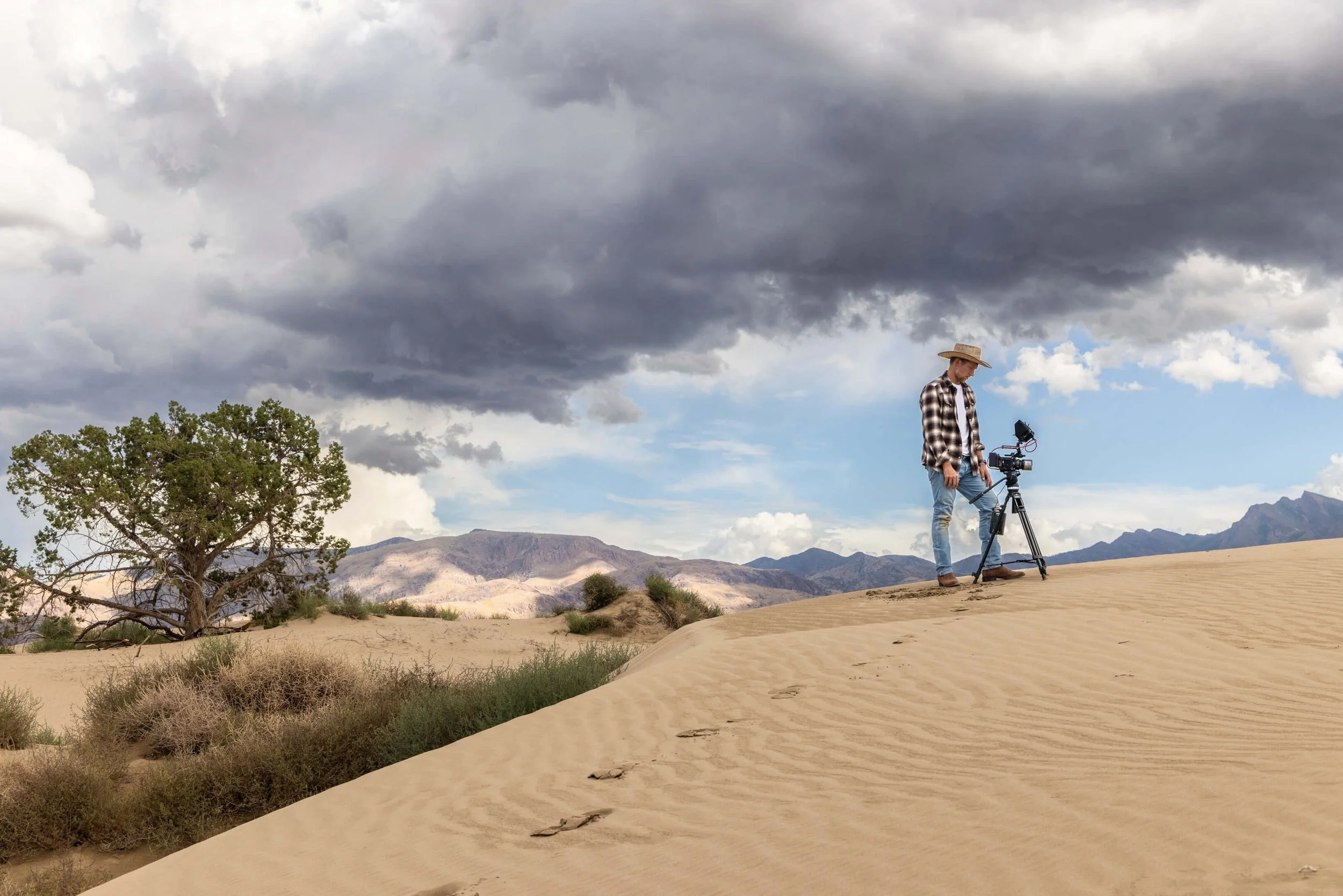 A man in plaid shirt and jeans operating a camera on tripod in a desert landscape with sand dunes, a tree, mountains in the distance, and dark storm clouds overhead