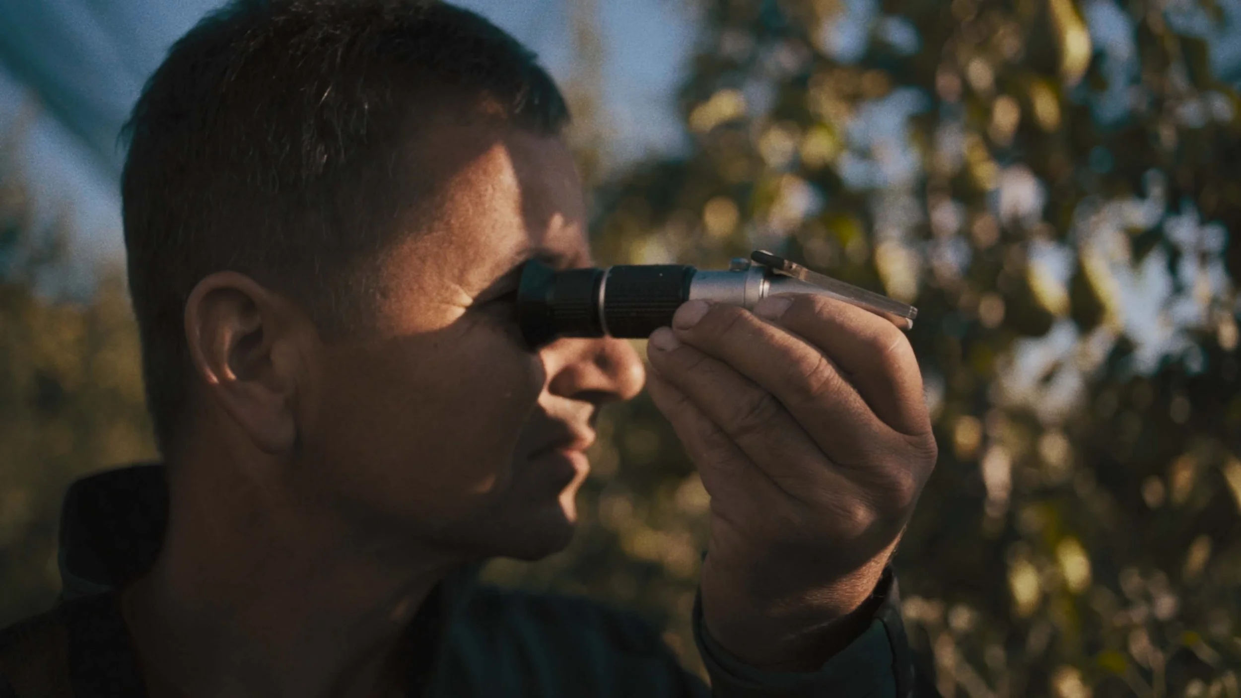 Person looking through binoculars outdoors with blurred background of trees and sunlight