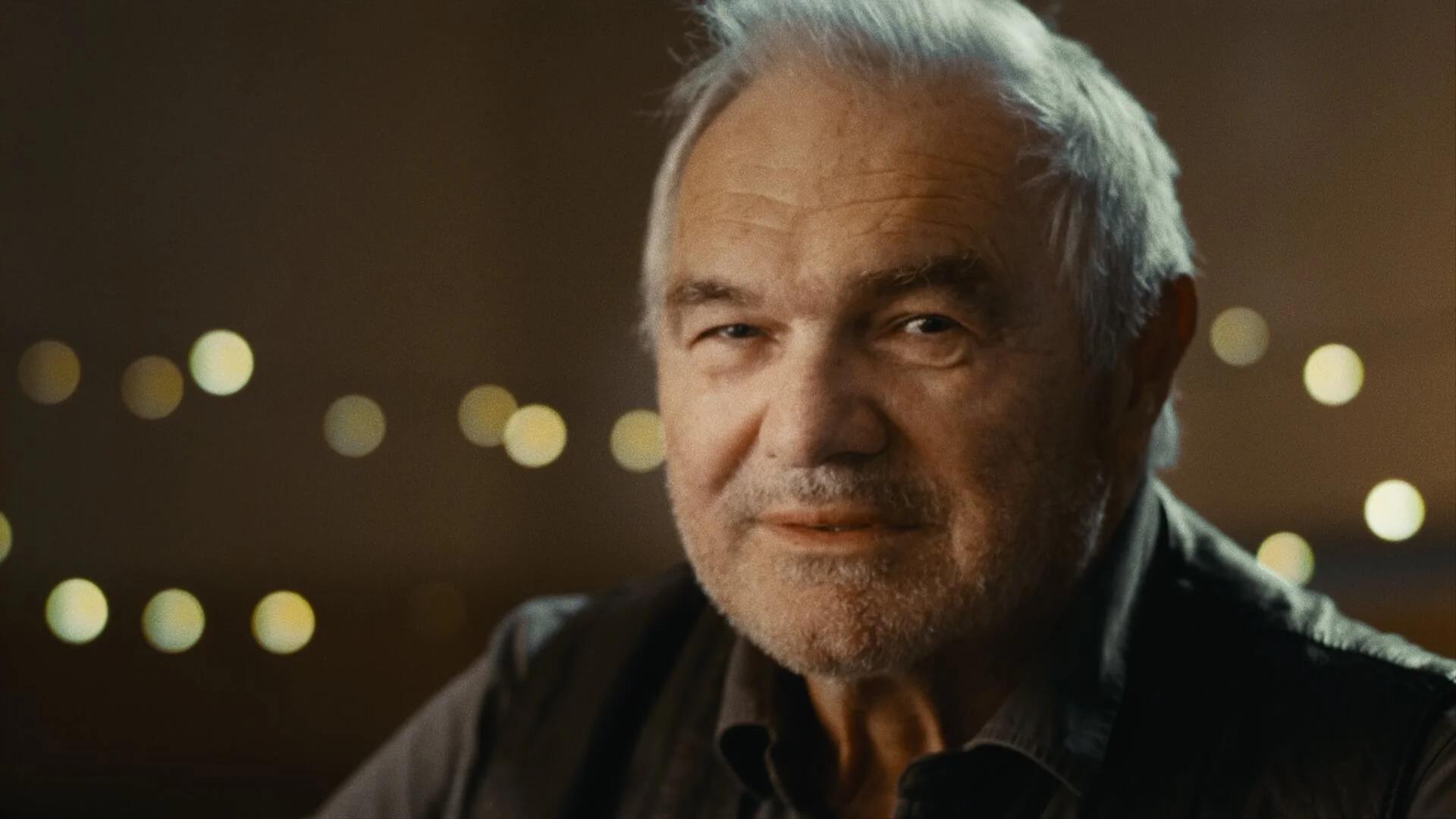 Close-up of an elderly man with gray hair and a beard, smiling softly, with out-of-focus lights in the background.