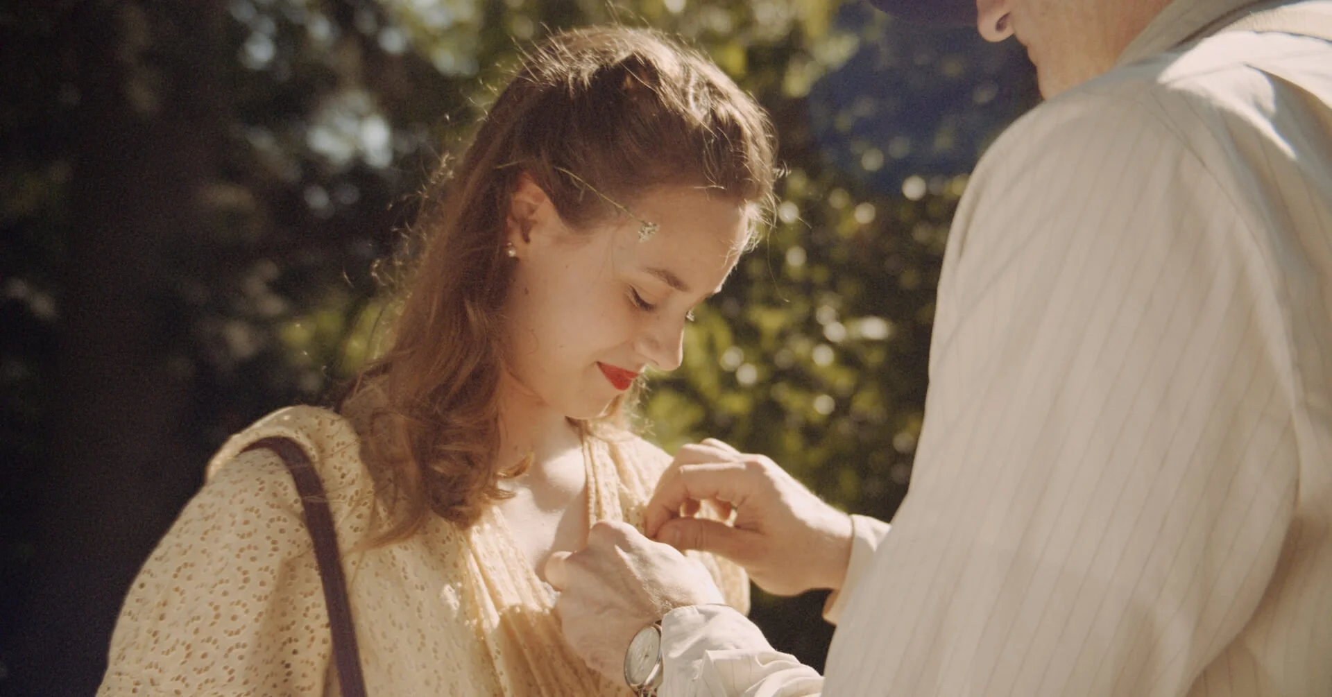 A woman with red lipstick and curly hair wearing a yellow dress is being pinned with a brooch by an older man in a striped shirt outdoors during the daytime.