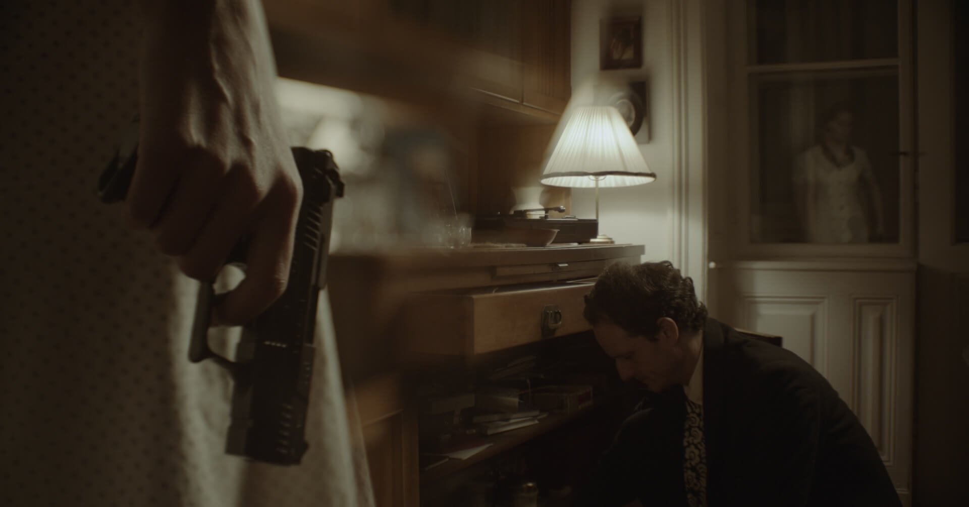 A woman is holding a gun while a man is kneeling on the floor in a dimly lit room, near a piano and a lamp.