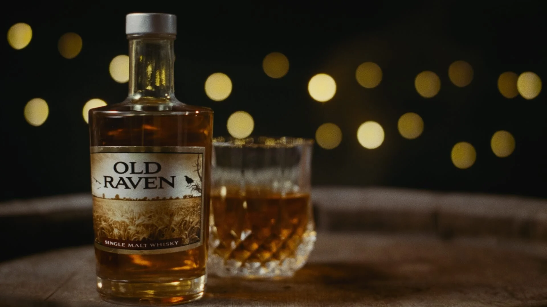 A bottle of Old Raven single malt whiskey and a glass of whiskey on a wooden surface with blurred warm yellow lights in the background.