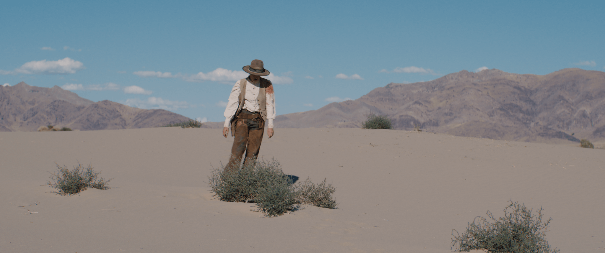 Person wearing vintage clothing walking in desert with mountains in the background under blue sky.