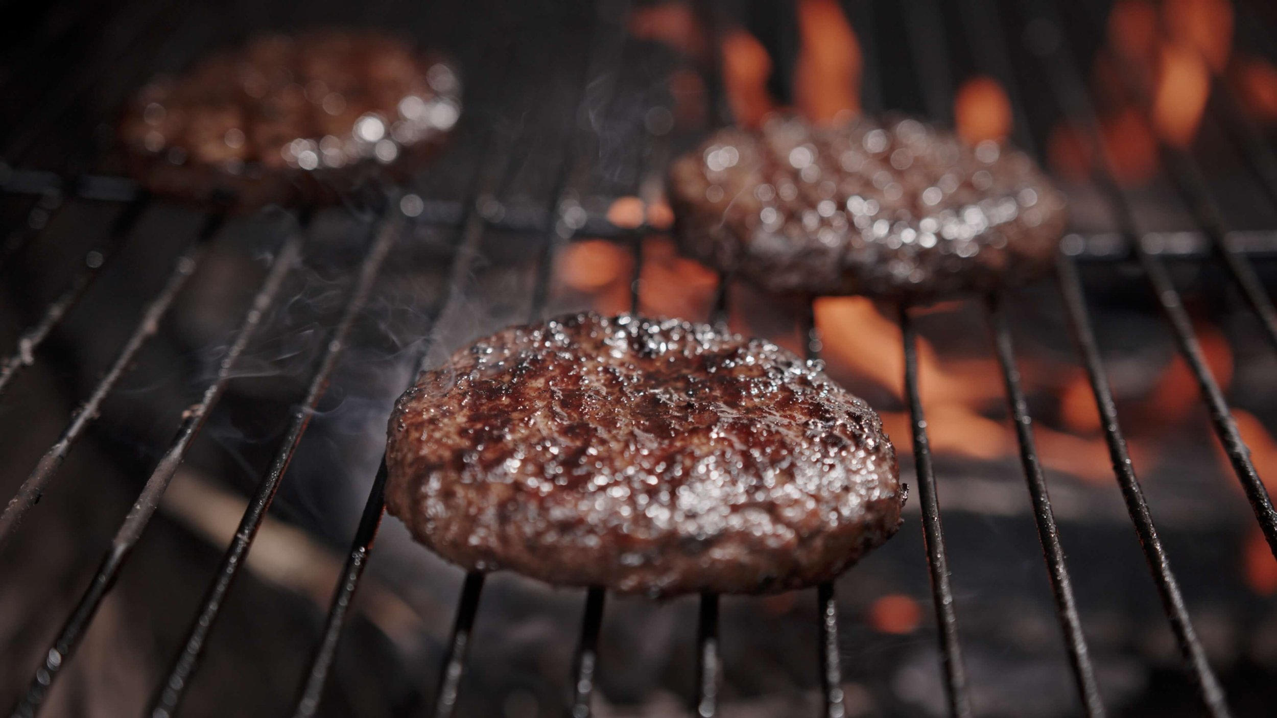 Hamburgers cooking on a barbecue grill with flames visible underneath.