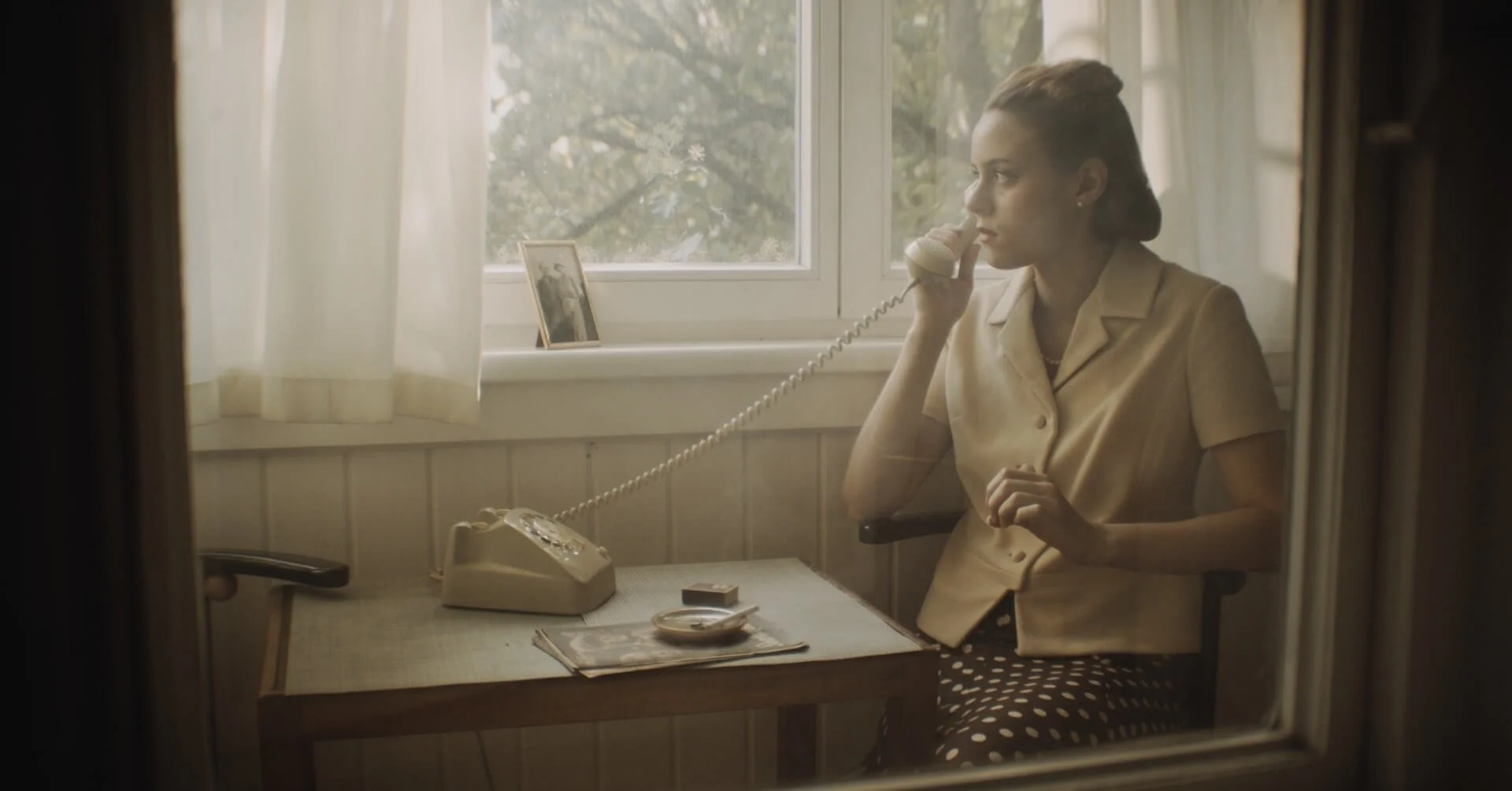 A woman with styled hair sitting at a desk, talking on a vintage rotary phone, with a window behind her showing trees outside, a framed photo on the windowsill, and a table with a magazine and a plate in a cozy, well-lit room.
