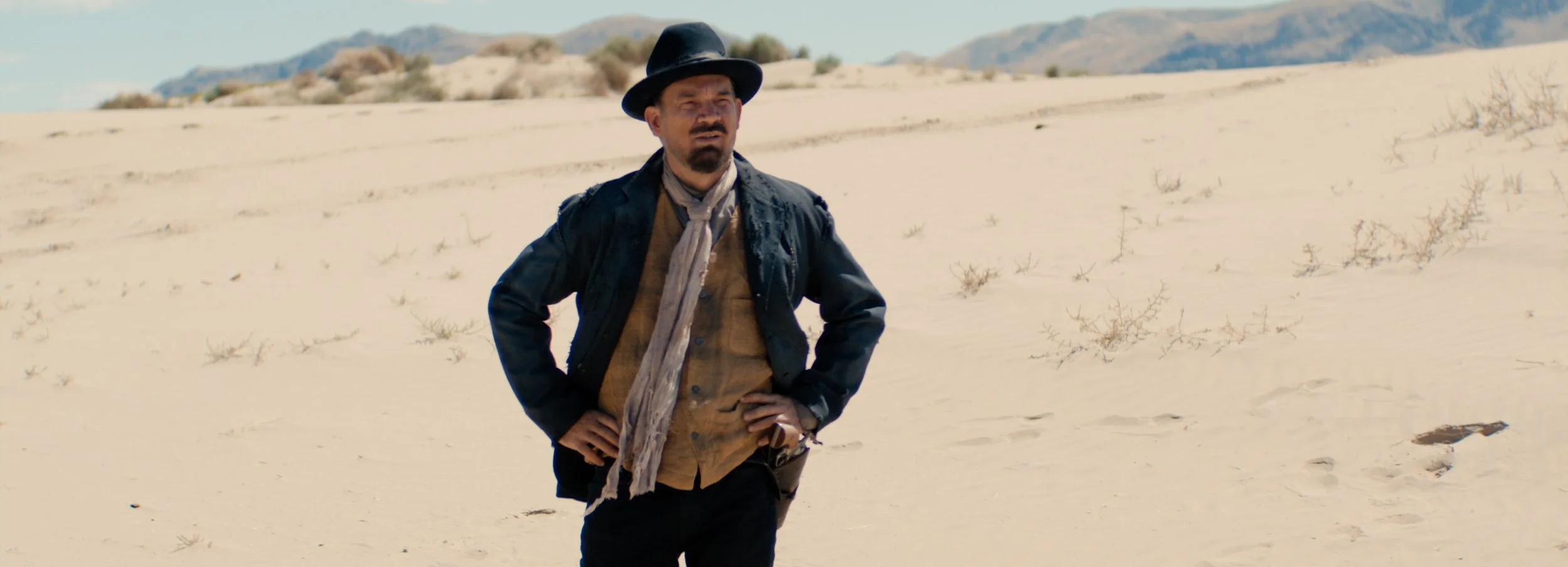 A man standing in a desert landscape with sand dunes and sparse vegetation, wearing a black hat, dark jacket, scarf, and brown shirt.
