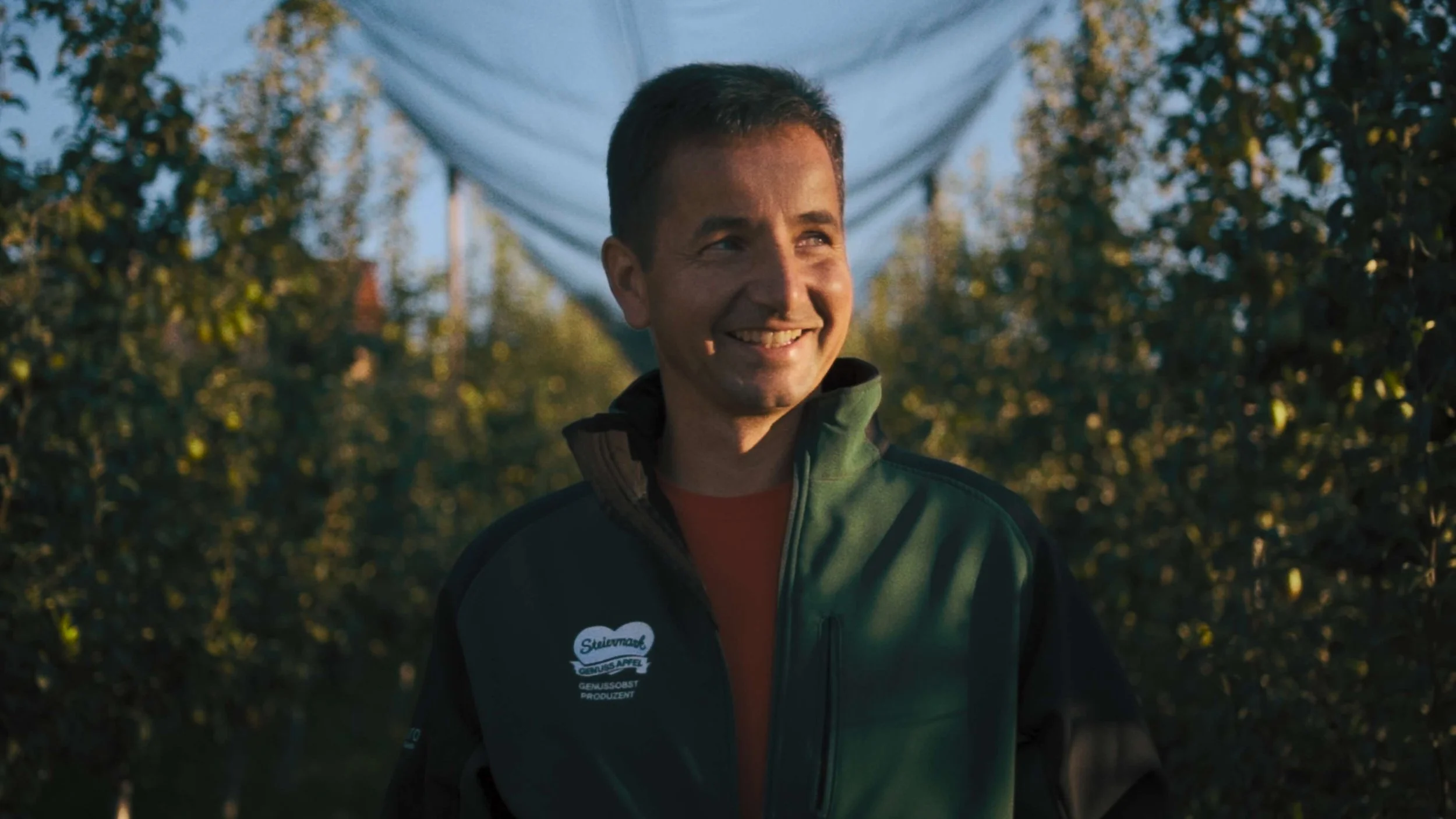 A man smiling outdoors in a green jacket under a white canopy, with trees and sunlight in the background.