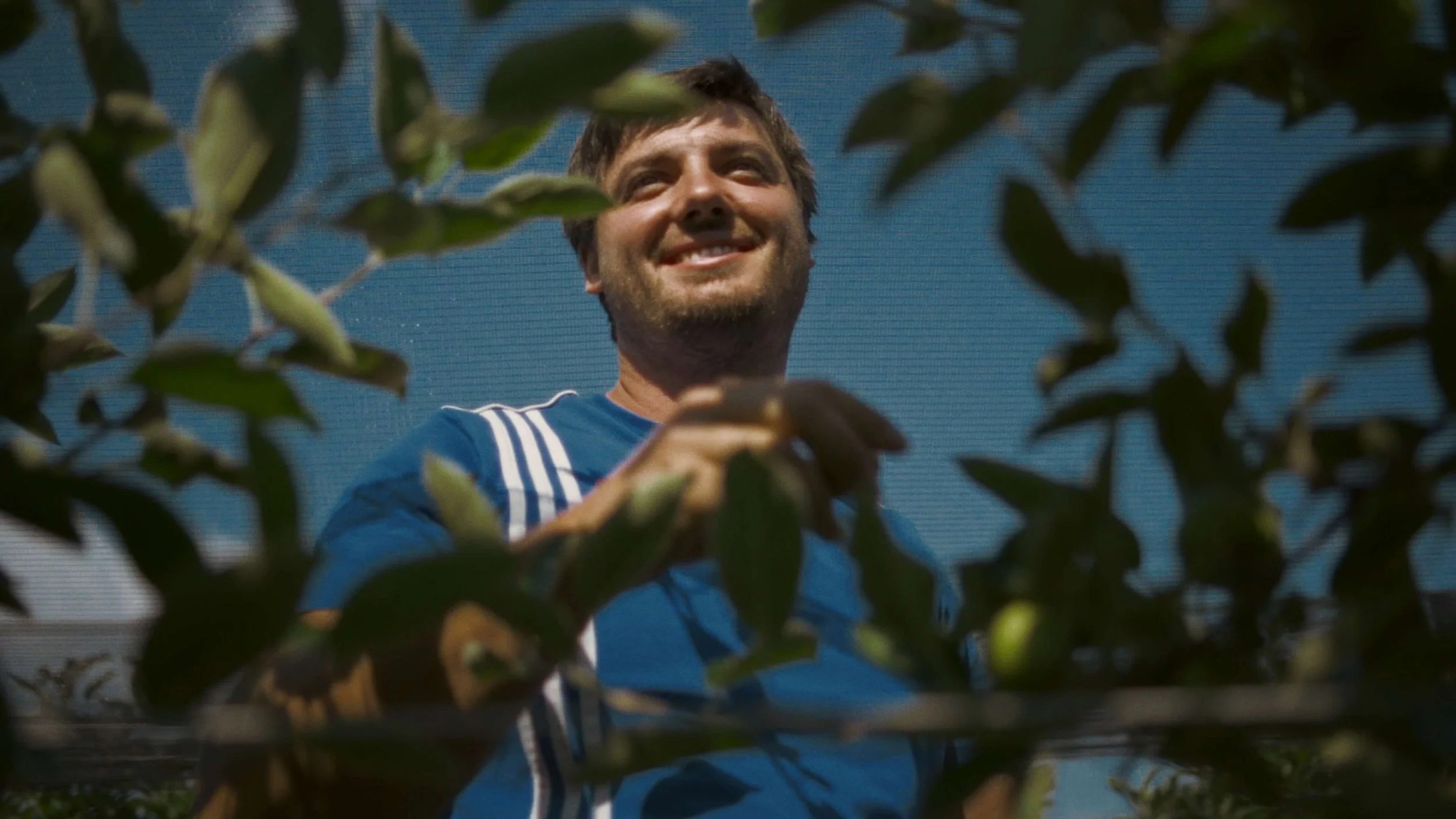A man smiling while working outdoors amidst green leaves, wearing a blue sports jersey.