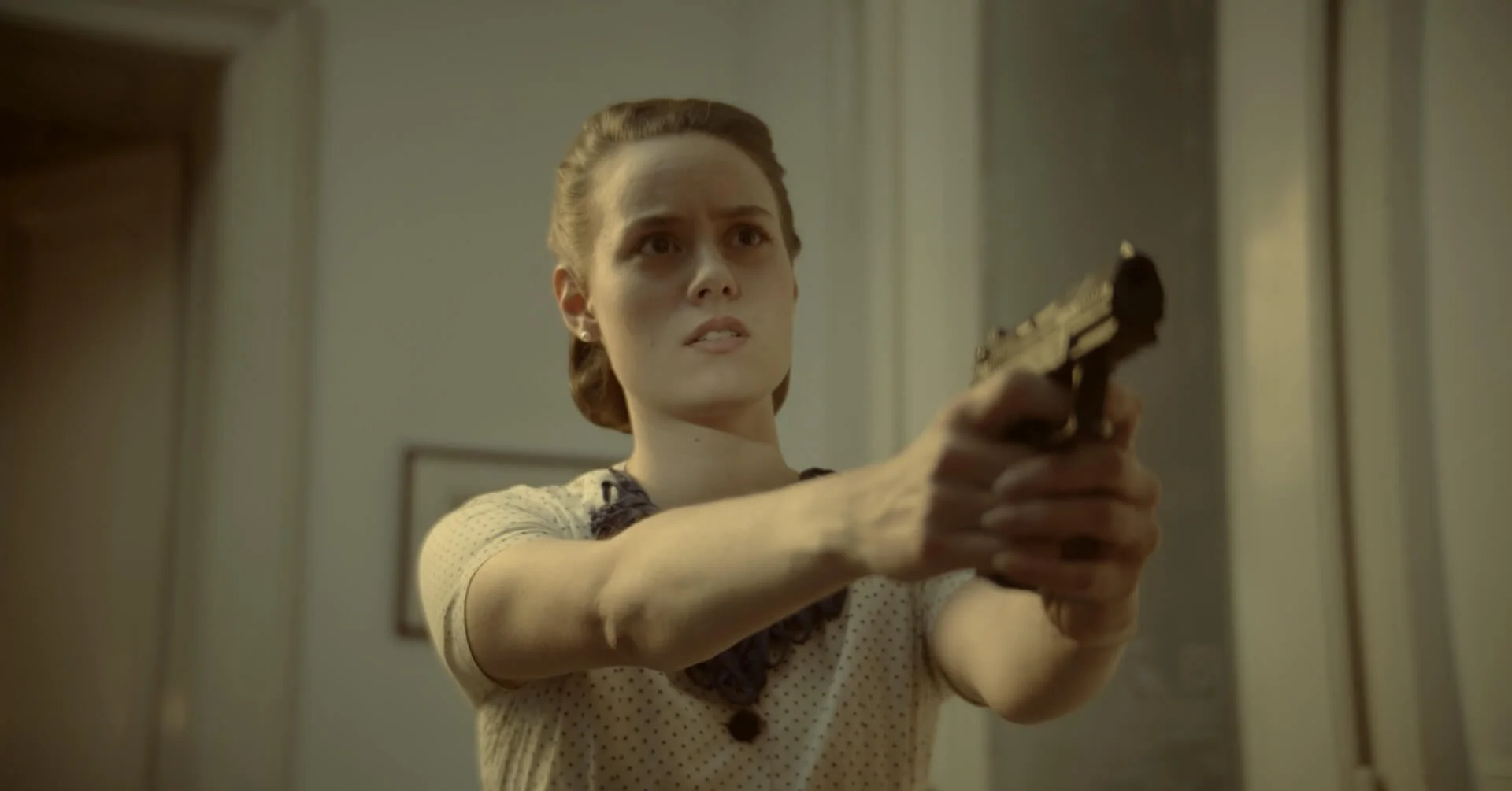 A woman with brown hair styled back, wearing a beige polka dot blouse with dark lace trim, is pointing a handgun with both hands, aiming at something off-screen. She has a serious and focused expression.