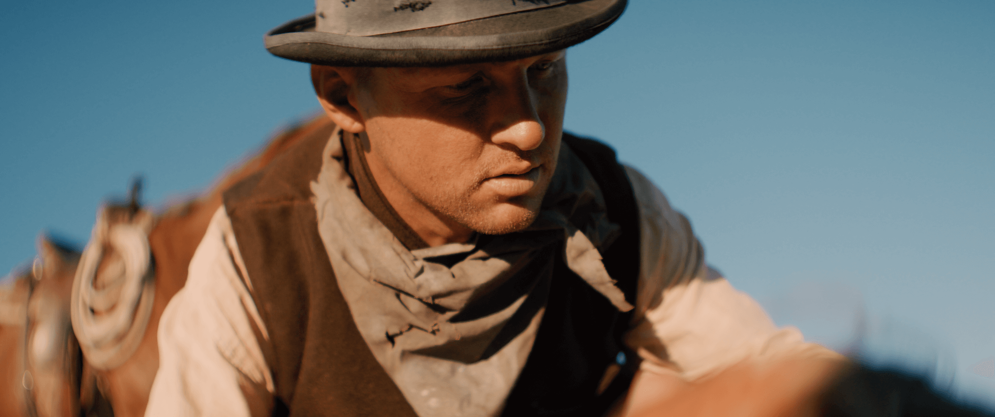 Close-up of a young man dressed as a cowboy, wearing a wide-brimmed hat, bandana, and vintage-style clothing, outdoors against a clear blue sky.