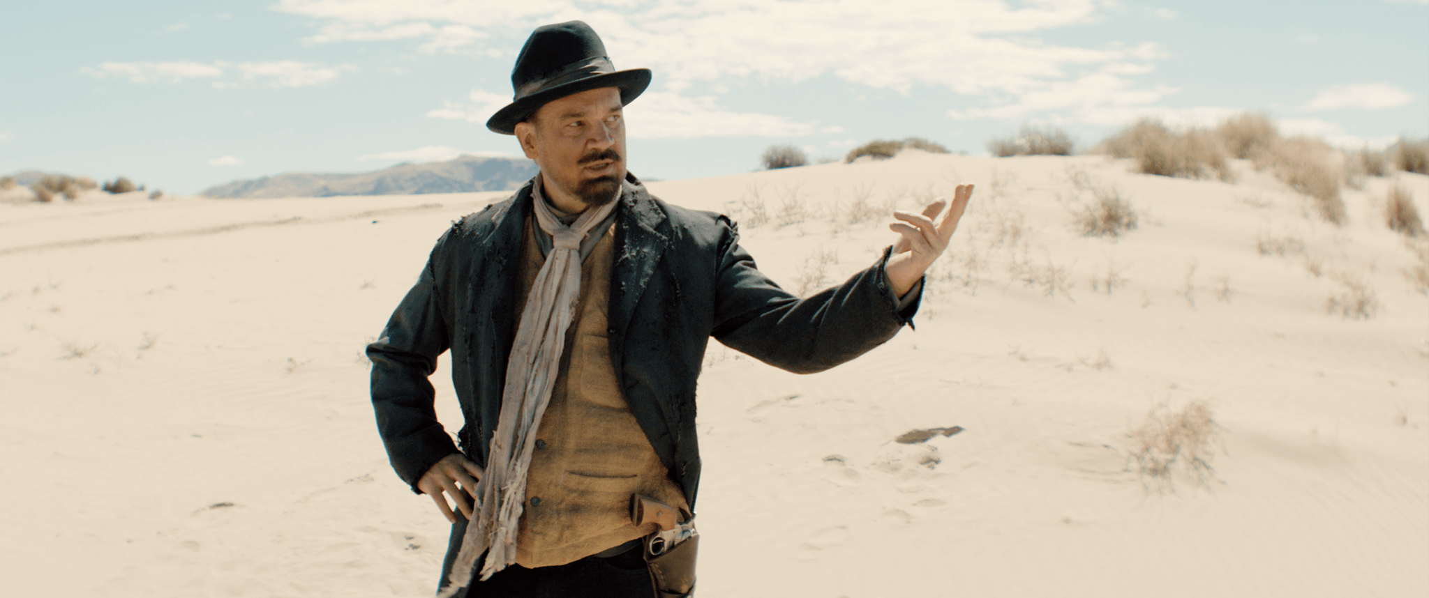 A man dressed in Old West style clothing, including a black hat, jacket, beige shirt, and scarf, stands in a desert landscape with sand dunes and sparse bushes, gesturing with his hand.