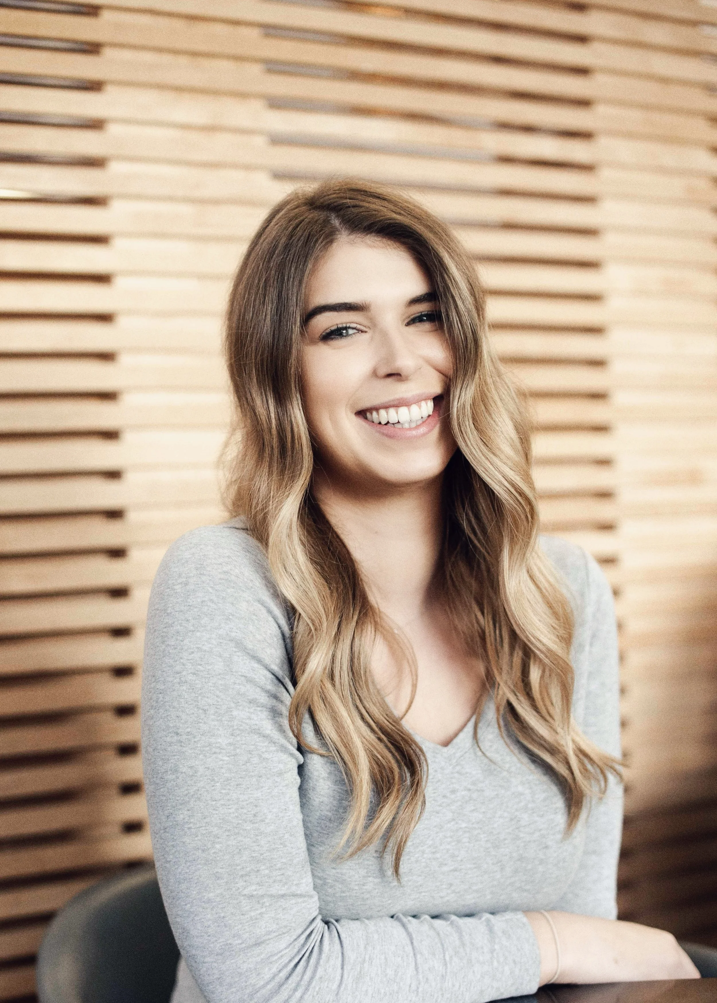 A woman smiling with long wavy hair, wearing a gray top, sitting in front of a wooden wall.