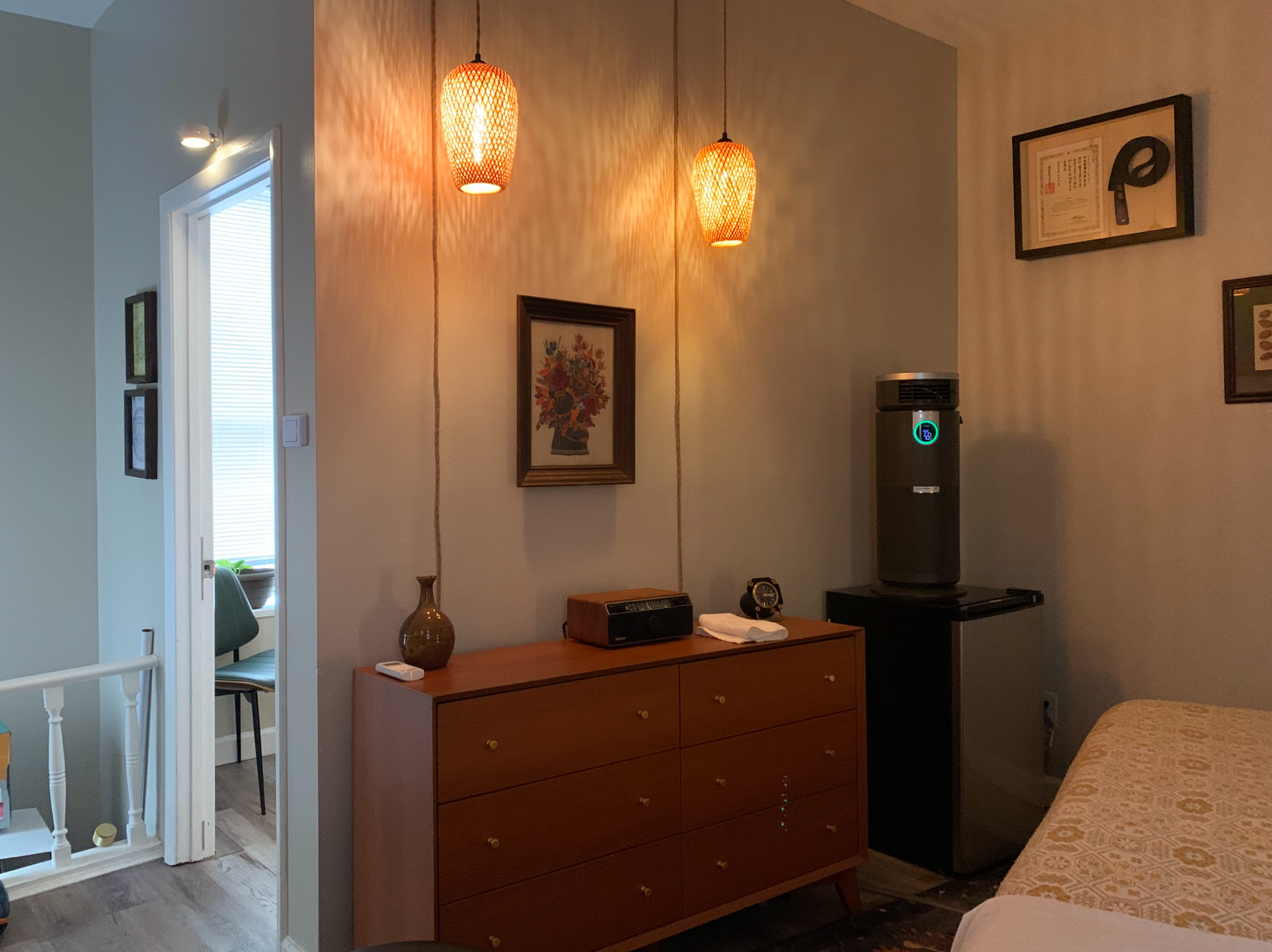 A cozy treatment room corner with a wooden dresser, a framed floral artwork, and two orange woven pendant lights hanging from the ceiling. Part of a table with a patterned blanket and a water cooler are visible.