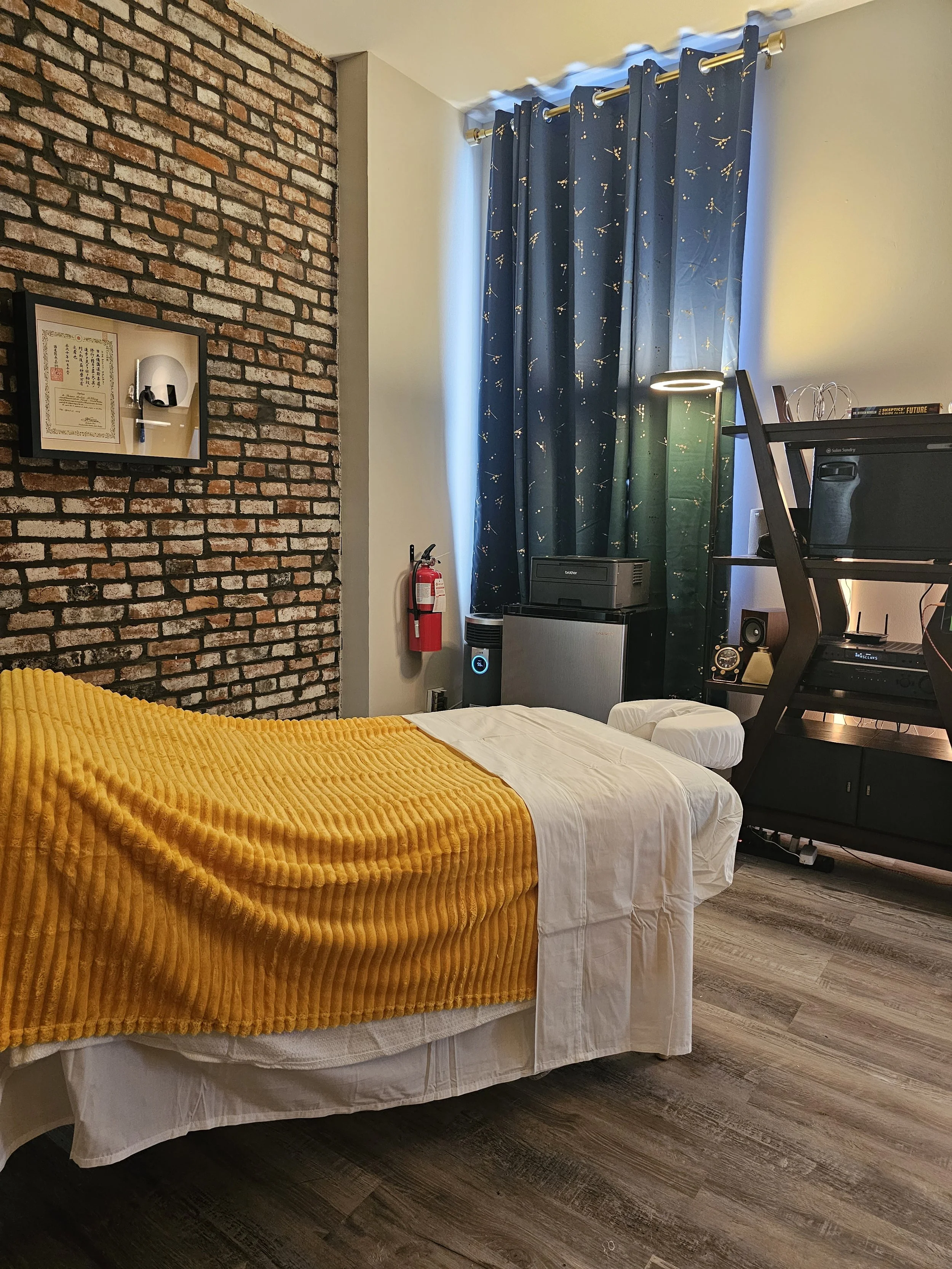 A massage bed with a golden yellow blanket in a room with a brick wall, a window with dark blue star-patterned curtains, and various electronic devices including a printer, a mini fridge, and a sound system.