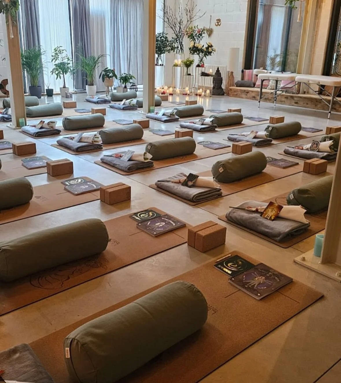 Yoga studio with mats laid out, including bolsters, blocks, blankets, and booklets. Plants and candles decorate the room, creating a calm atmosphere.