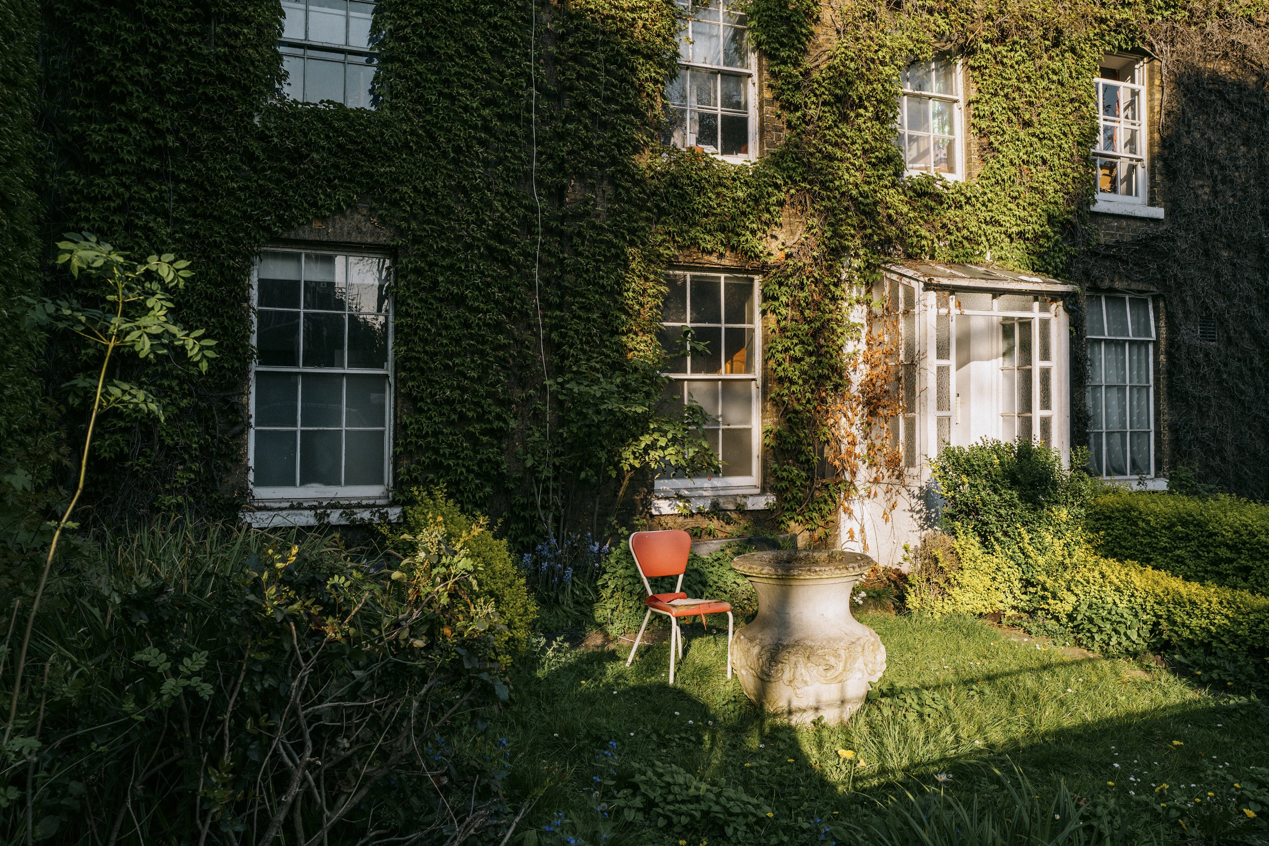 An ivy-covered house with multiple white-framed windows, a small garden with bushes and flowers, and a red chair situated next to an ornate stone planter.