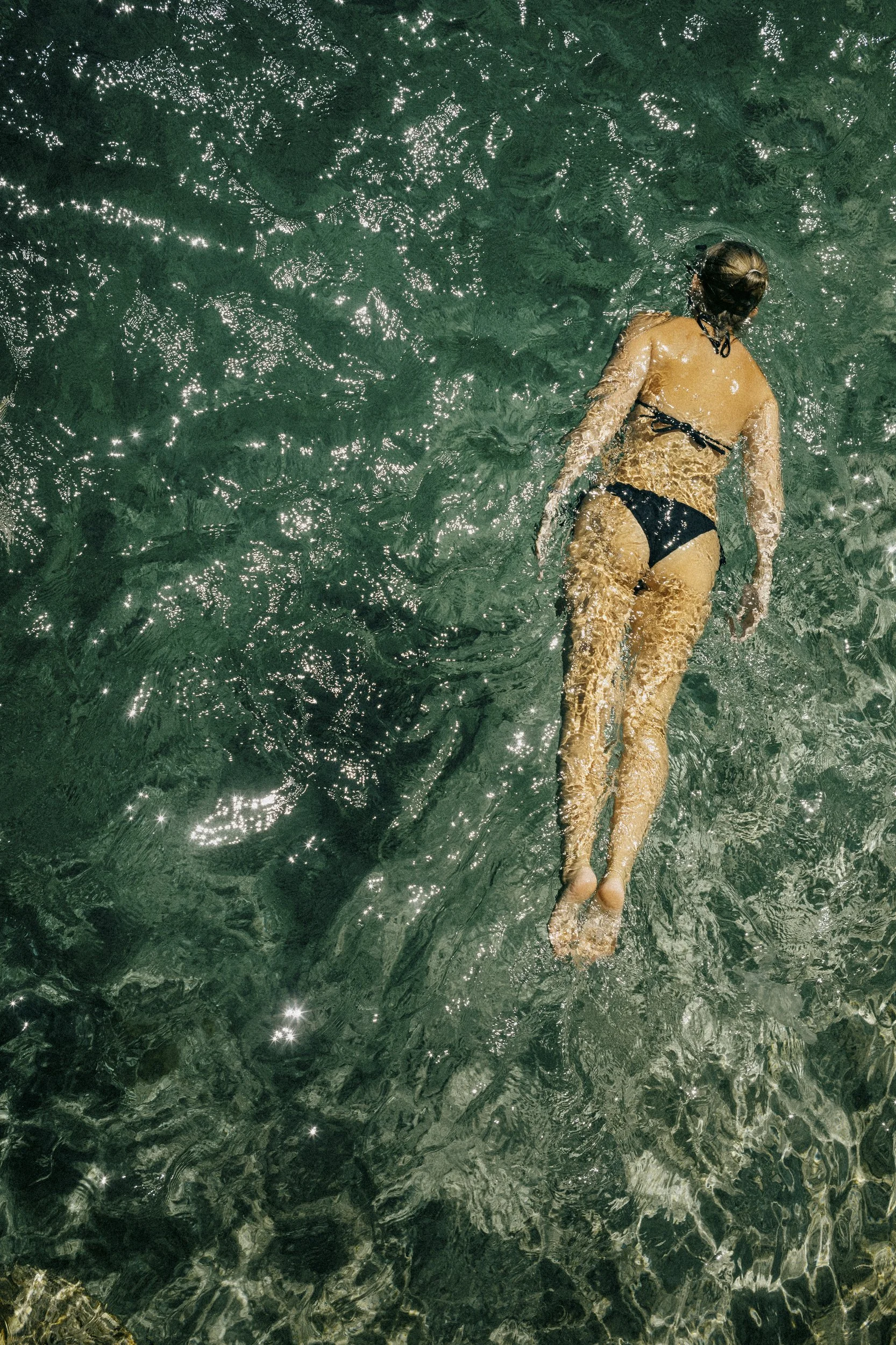 A person floating on their back in clear, green water, wearing a black bikini and sunglasses.