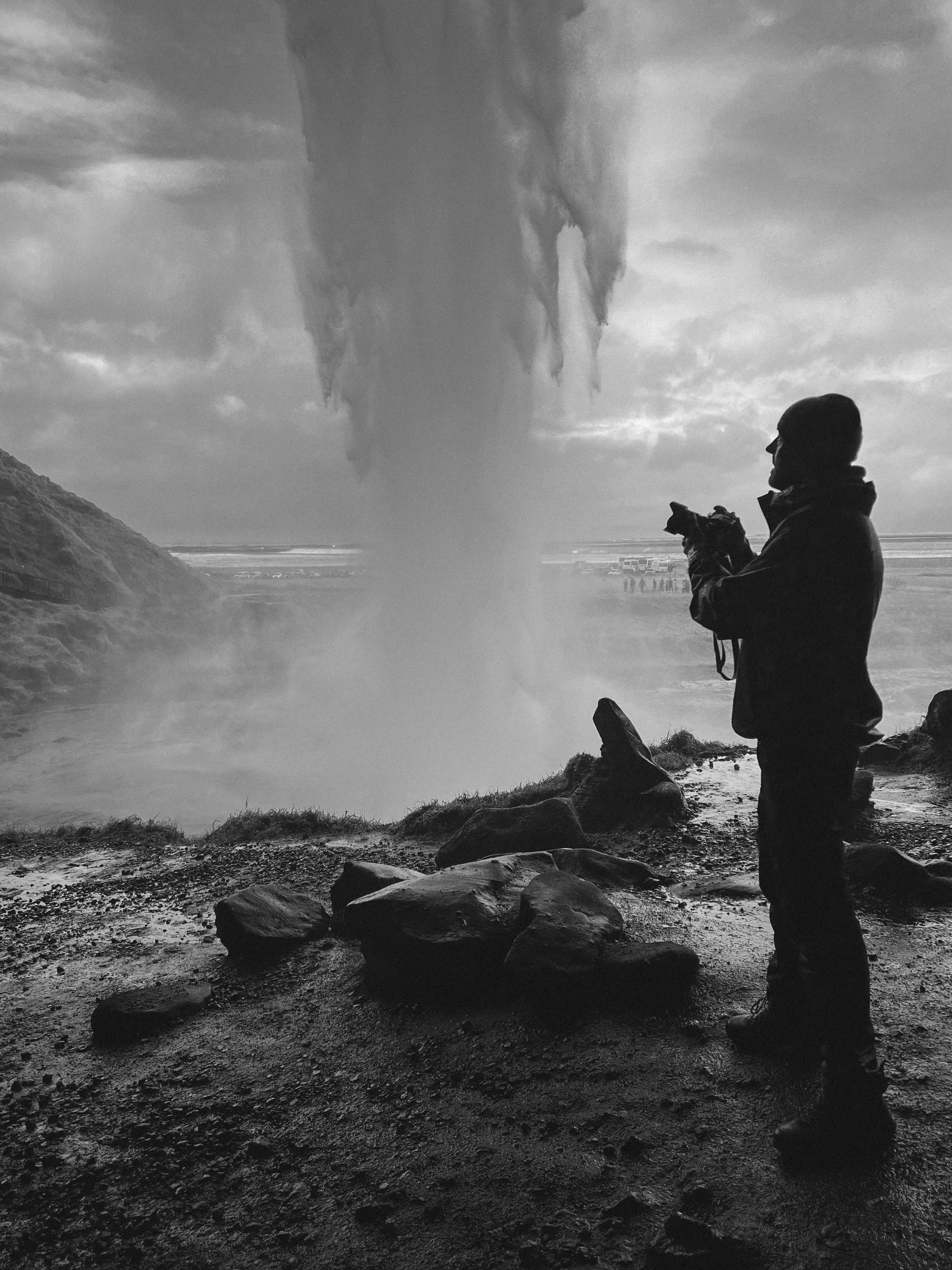 A person stands on rocky ground, taking a photo with a camera, overlooking a large geyser erupting in a volcanic landscape during daytime.