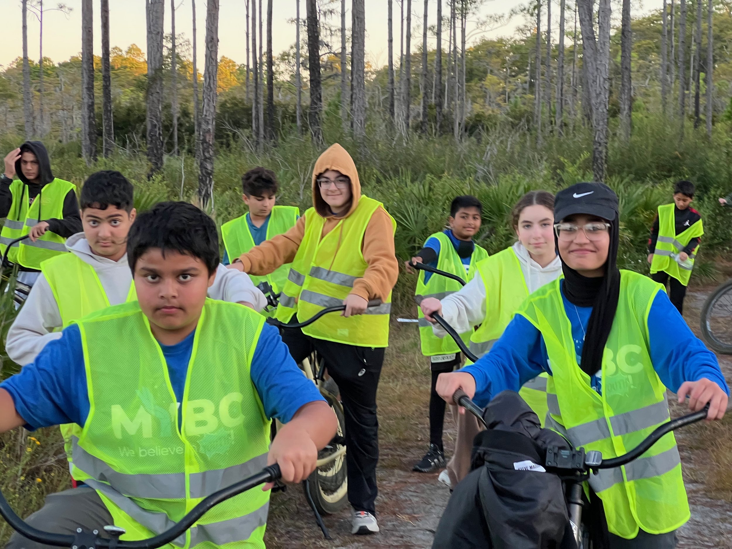 A group of children wearing bright yellow safety vests with the letters 'MBC' on them, holding bicycles, standing on a dirt trail in a forested area with tall trees and green foliage.