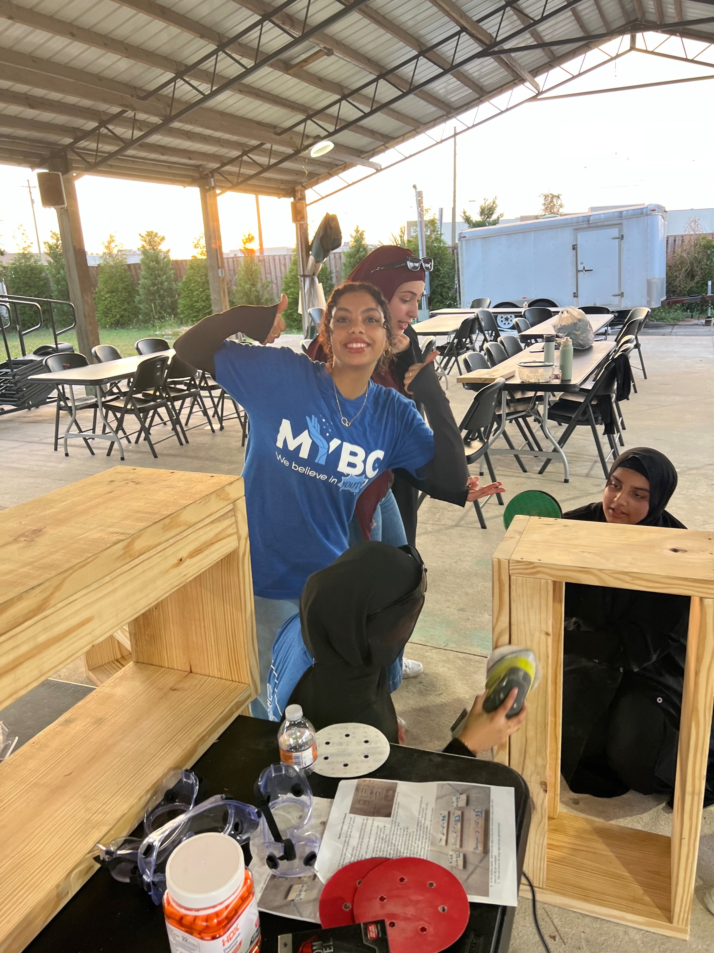 Group of young women at an outdoor or semi-outdoor table setup with construction materials, sunglasses, and a water bottle, smiling and engaging in conversation during sunset.