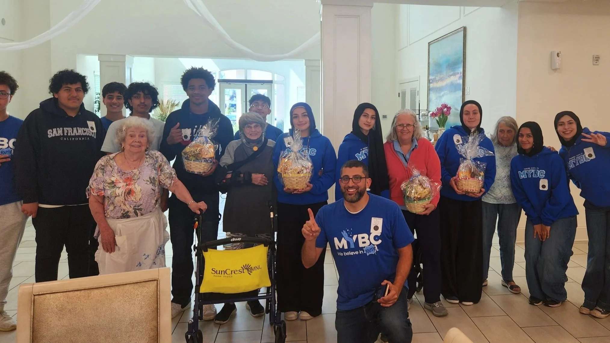 Group of diverse people, including elderly women, young adults, and teens, gathered in a lobby or reception area, smiling and holding gift baskets wrapped in cellophane. Some individuals are wearing blue shirts with 'MYBC' on them, indicating a community or youth organization. The setting is bright and welcoming, with artwork and on-site decor visible.