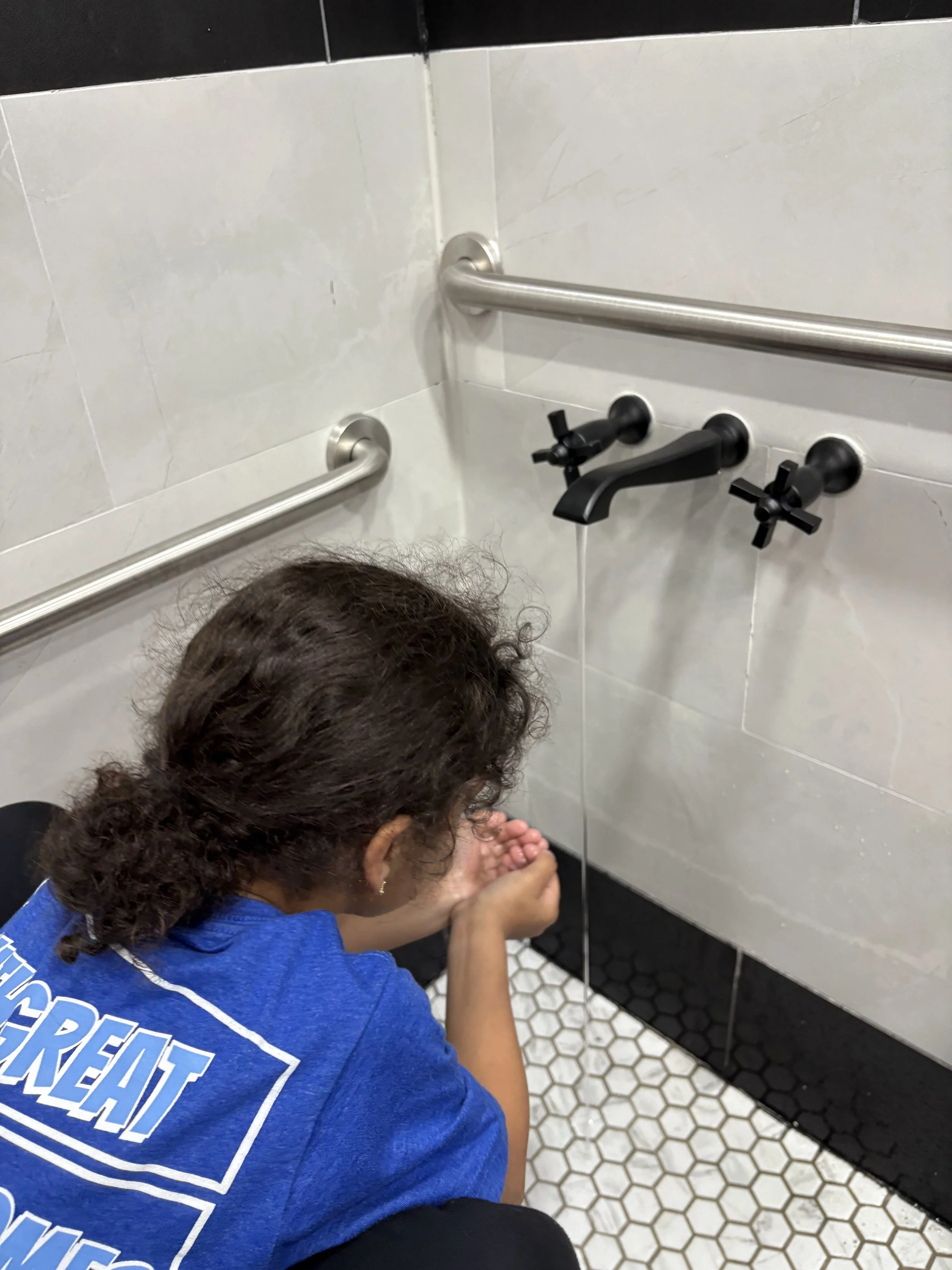 A woman in a blue shirt washing her hands under a faucet in a bathroom with white tiled walls and hexagonal floor tiles.