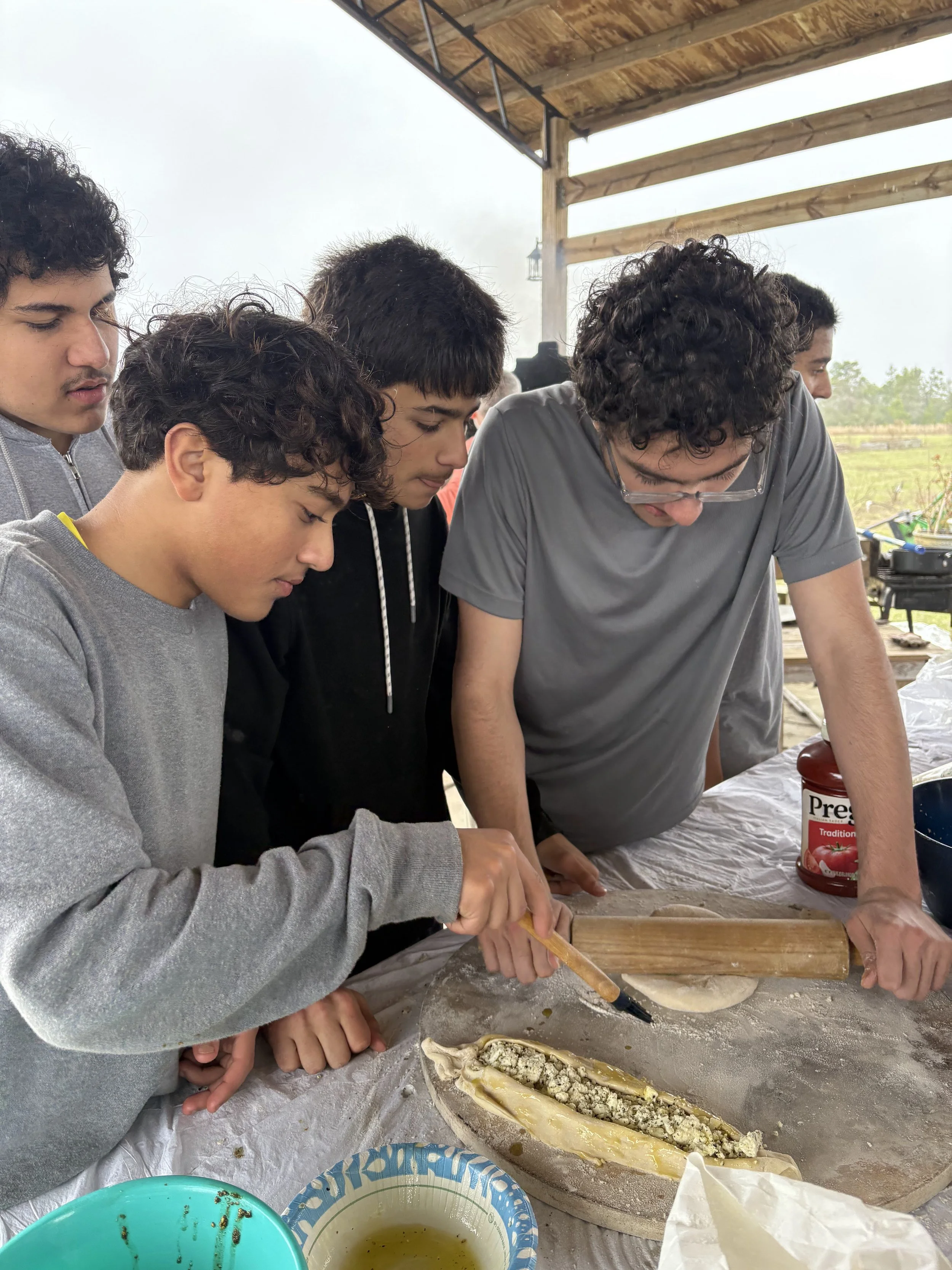 A group of five young people preparing a long piece of dough filled with cheese and other ingredients under a wooden roofed outdoor shelter, with one person rolling and another cutting the dough.