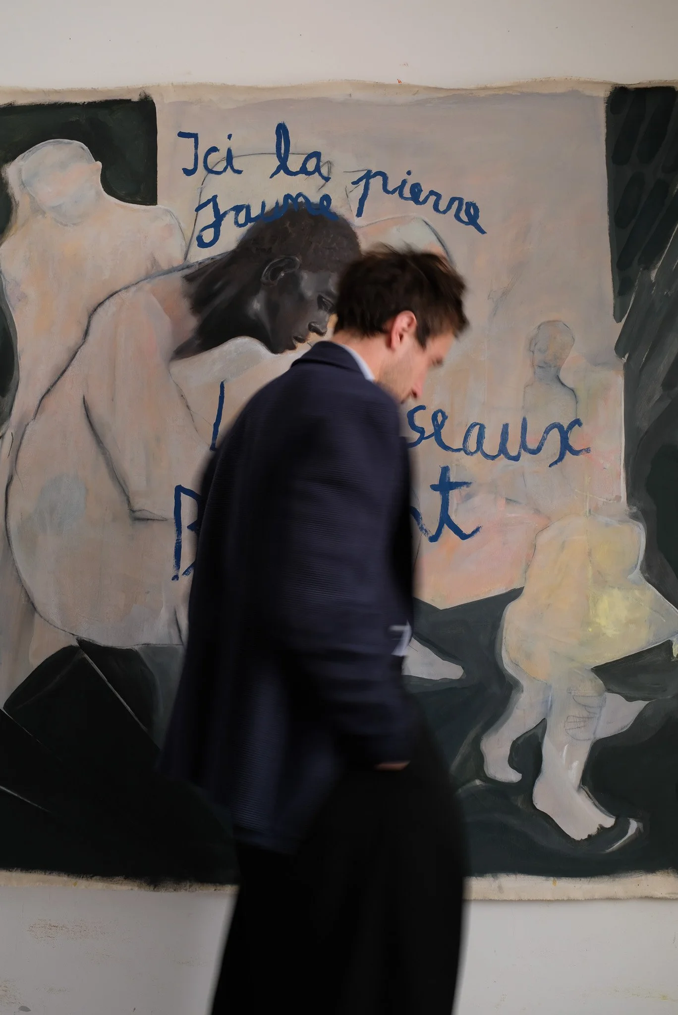 A young man walking past a mural with abstract and sketch-like human figures and French writing.